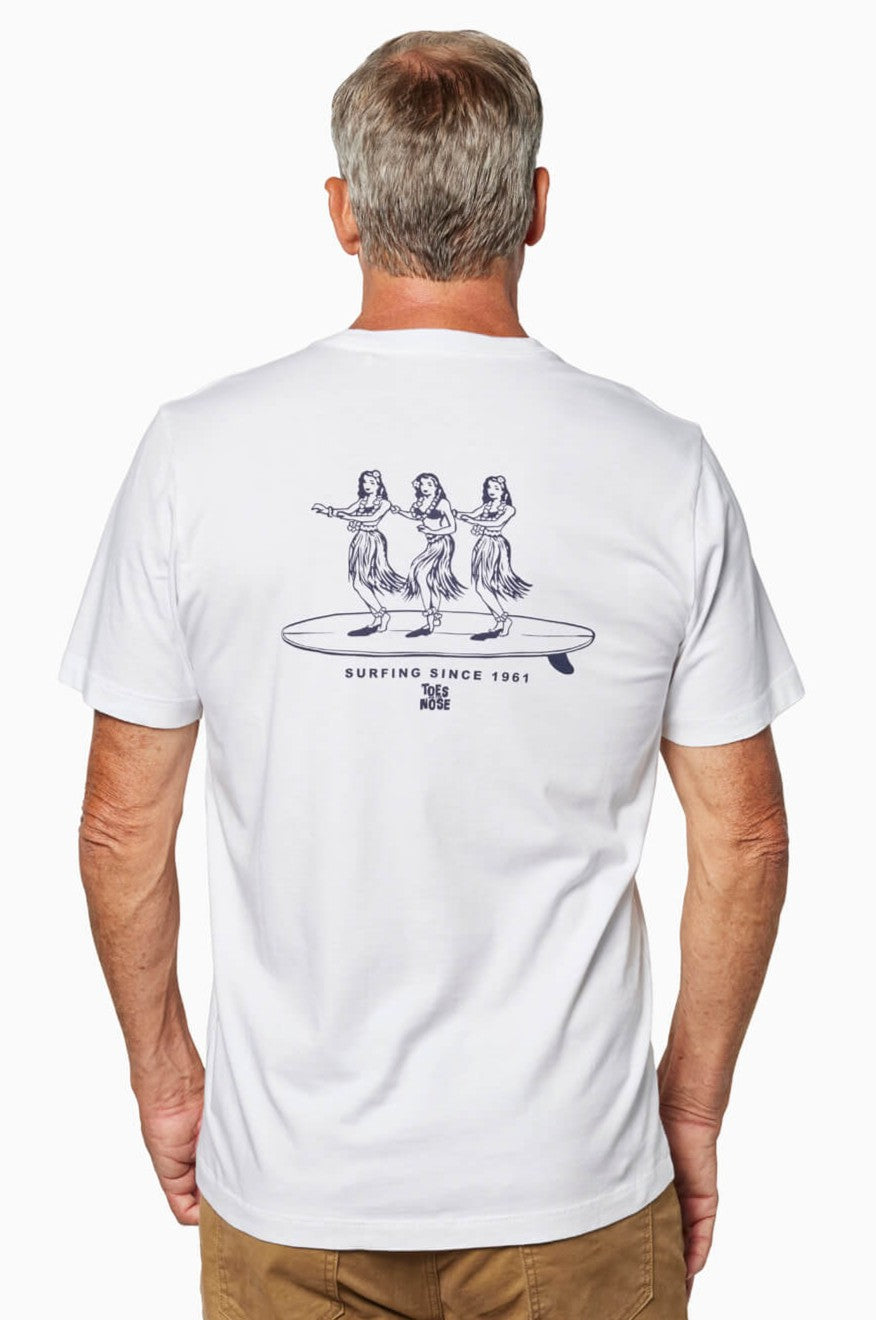 A man with gray hair wears the Toes on the Nose Hula Board Short Sleeve Tee, a relaxed fit white T-shirt featuring three hula dancers on a surfboard and “Surfing Since 1961” text. Crafted from premium Peruvian cotton; hand wash cold recommended.