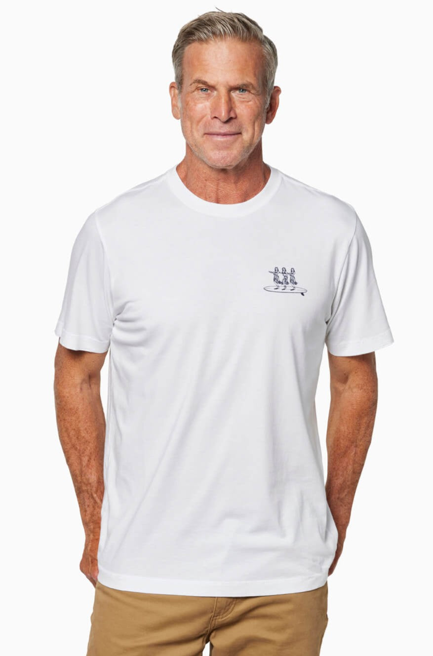 A middle-aged man stands smiling with his hands behind his back, wearing the Toes on the Nose Hula Board Short Sleeve Tee in white—made from premium Peruvian cotton with a small blue chest graphic—and khaki pants, against a white background.