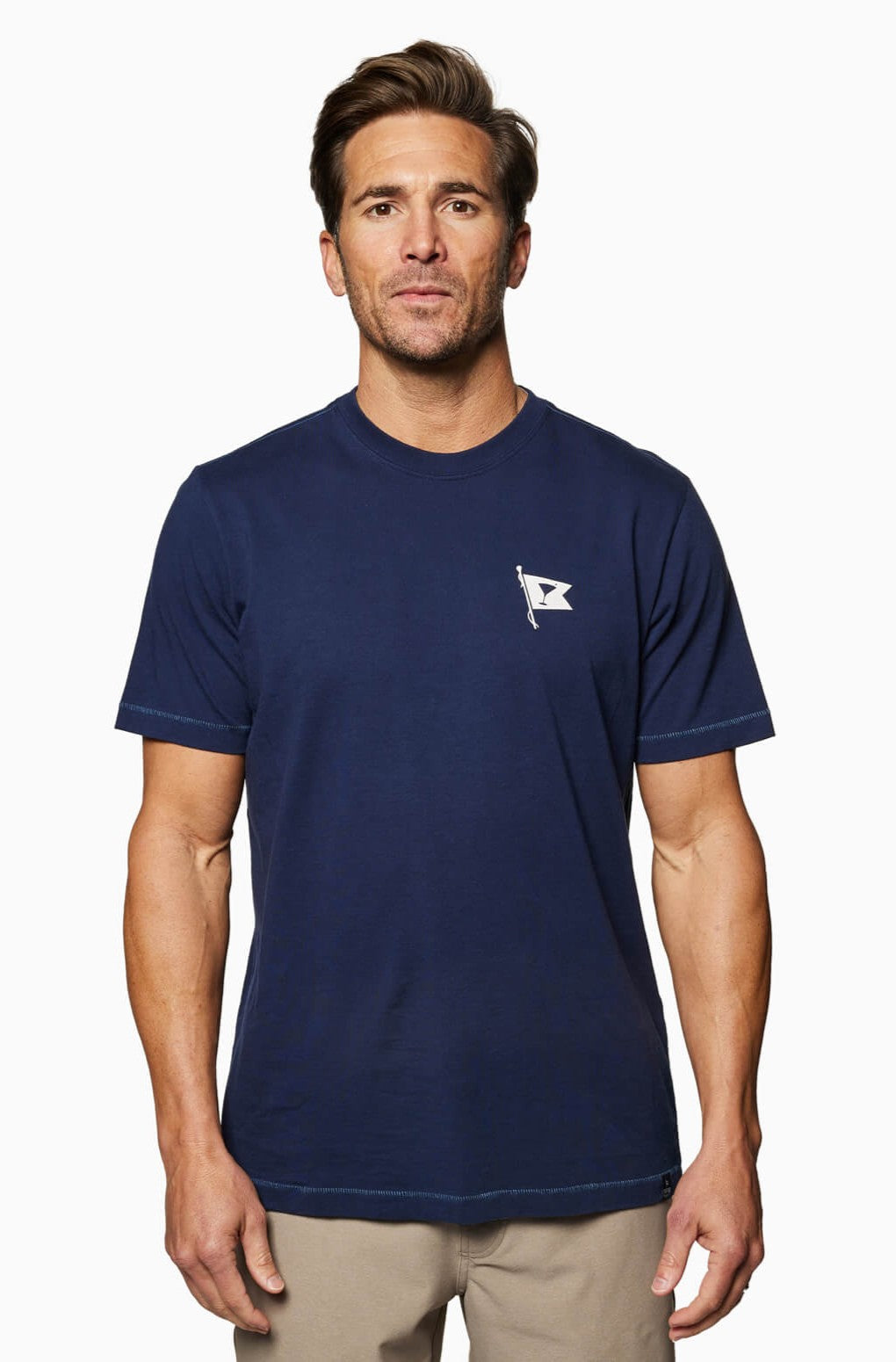 A man faces forward wearing the Toes on the Nose Yacht Club Short Sleeve Tee in navy, featuring a small white flag logo on the left chest, paired with beige pants, set against a plain white background.