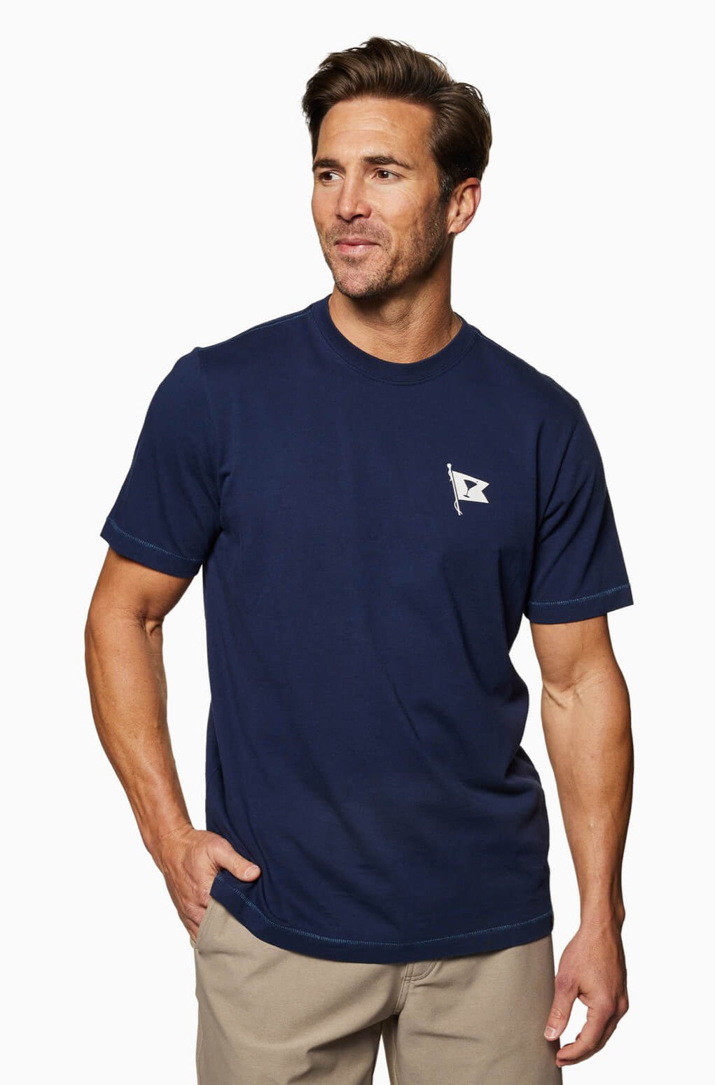 A man smiles to the side, wearing the Toes on the Nose Yacht Club Short Sleeve Tee in navy Peruvian cotton with a small white flag chest logo, paired with beige pants against a white background and one hand in his pocket.
