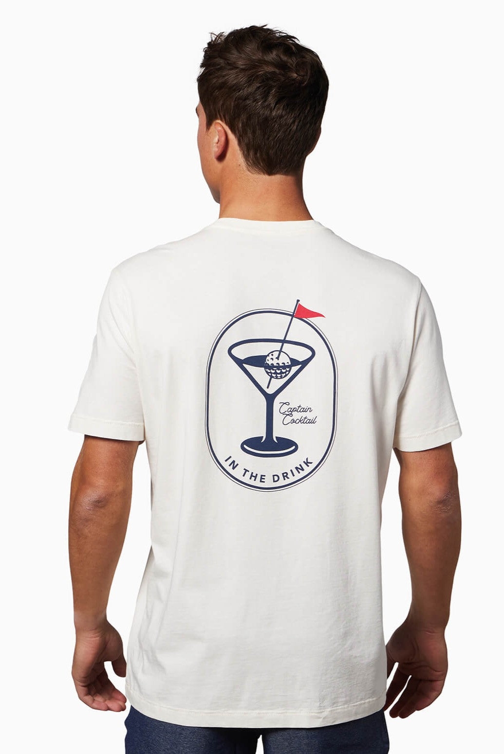 A man wearing the Toes on the Nose In The Drink Short Sleeve Tee, a white premium Peruvian cotton shirt with a back graphic of a martini glass holding a golf ball and red flag, plus the text: Transfusion Cocktail, Captain Cocktail, and IN THE DRINK.