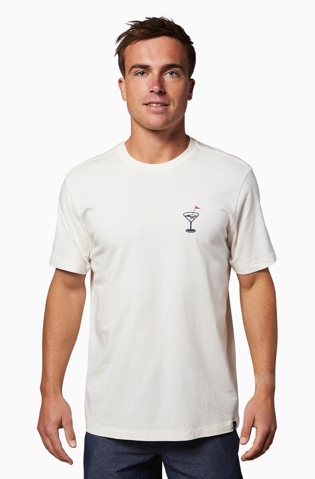 A man wearing the Toes on the Nose "In The Drink" Short Sleeve Tee in White Multi, featuring a small cocktail glass graphic on the left chest, stands and smiles against a white background.