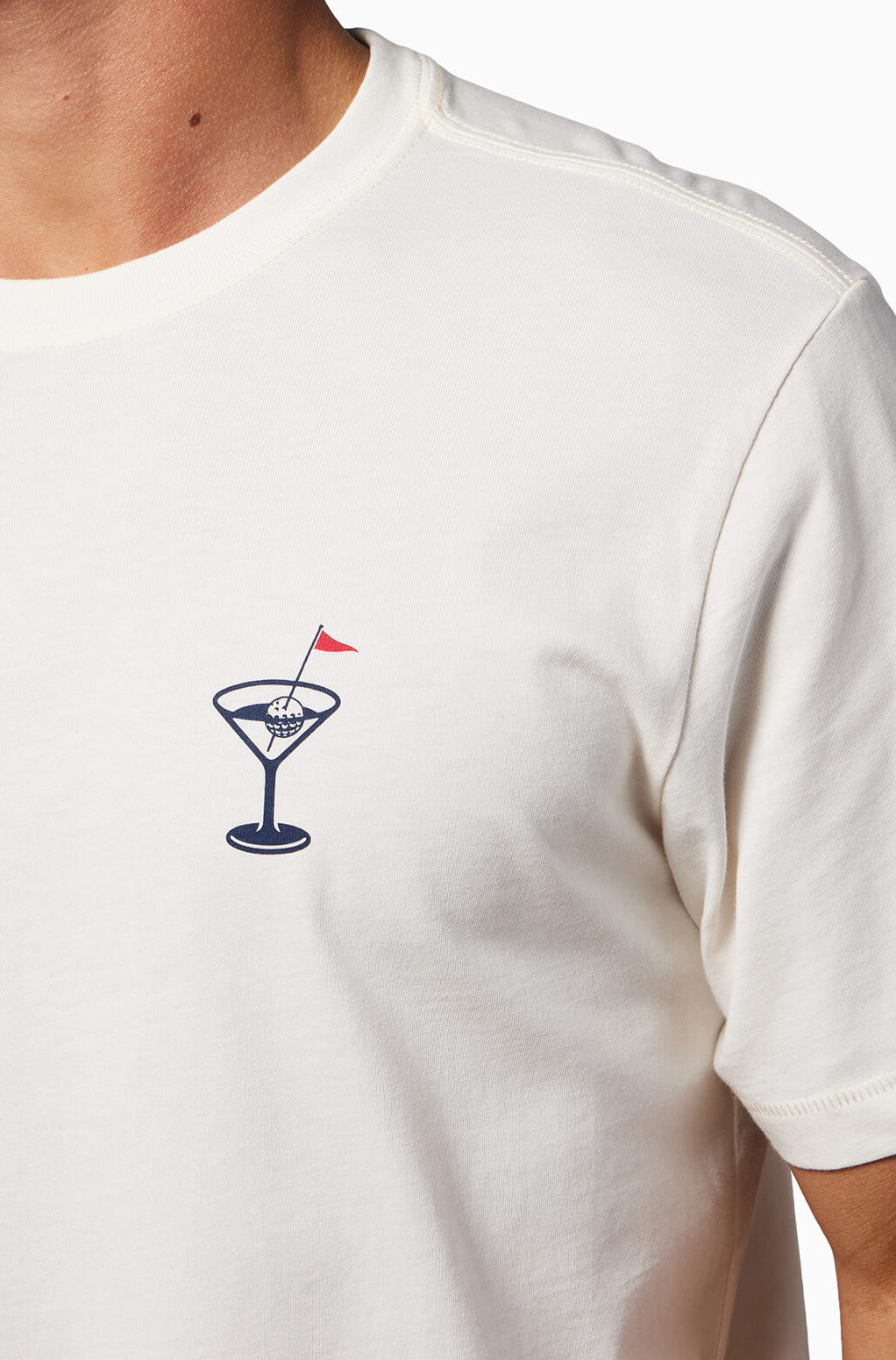 A person wears the Toes on the Nose In The Drink Short Sleeve Tee, an off-white premium Peruvian cotton shirt with a small cocktail glass, globe, and red flag graphic on the left chest.