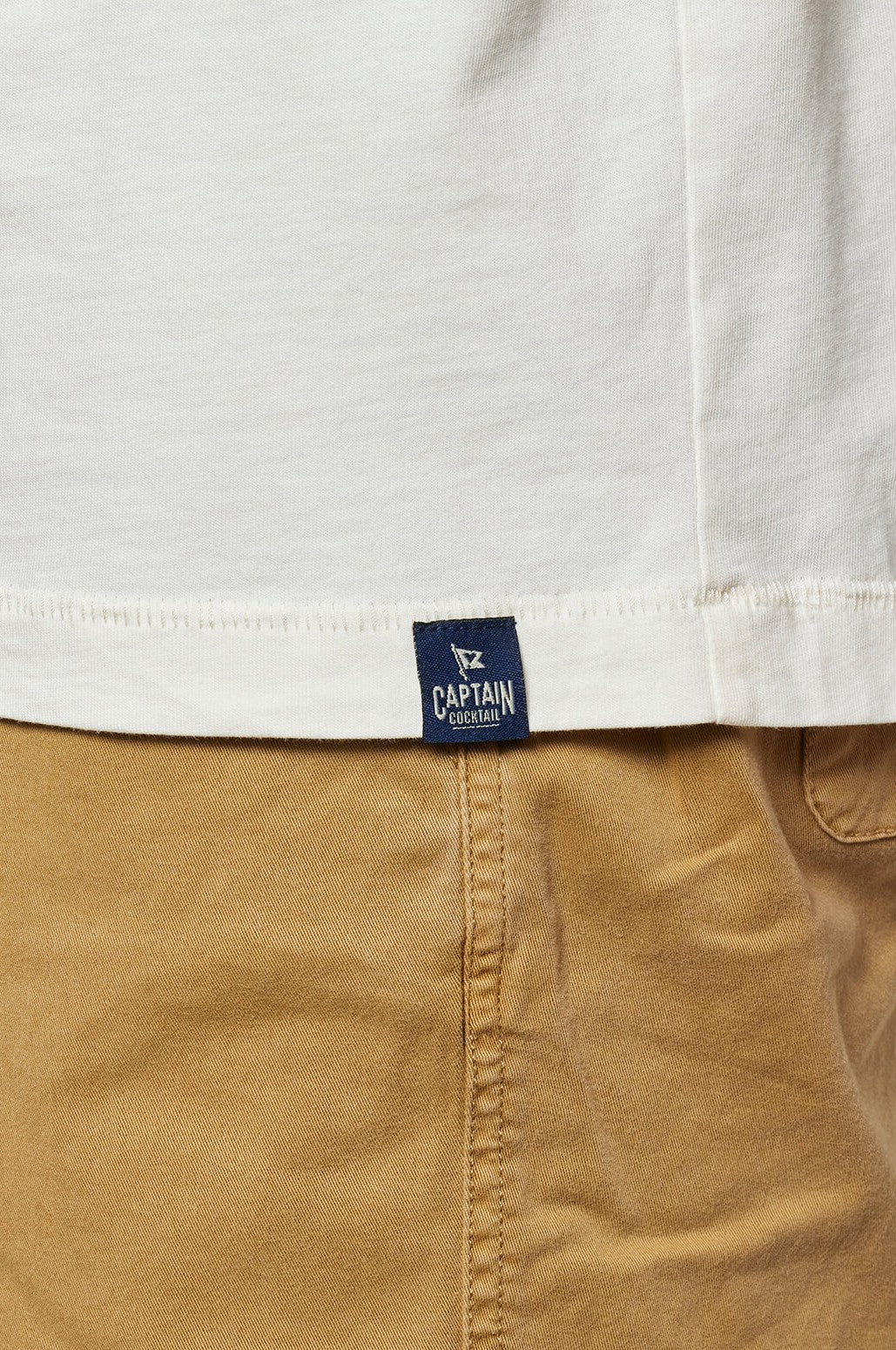 Close-up of a person in tan pants and a white Beers And Bogeys shirt by Toes on the Nose, crafted from Premium Peruvian Cotton with a relaxed fit and a small navy tag near the hem reading CAPTAIN CENTAUR above its logo.