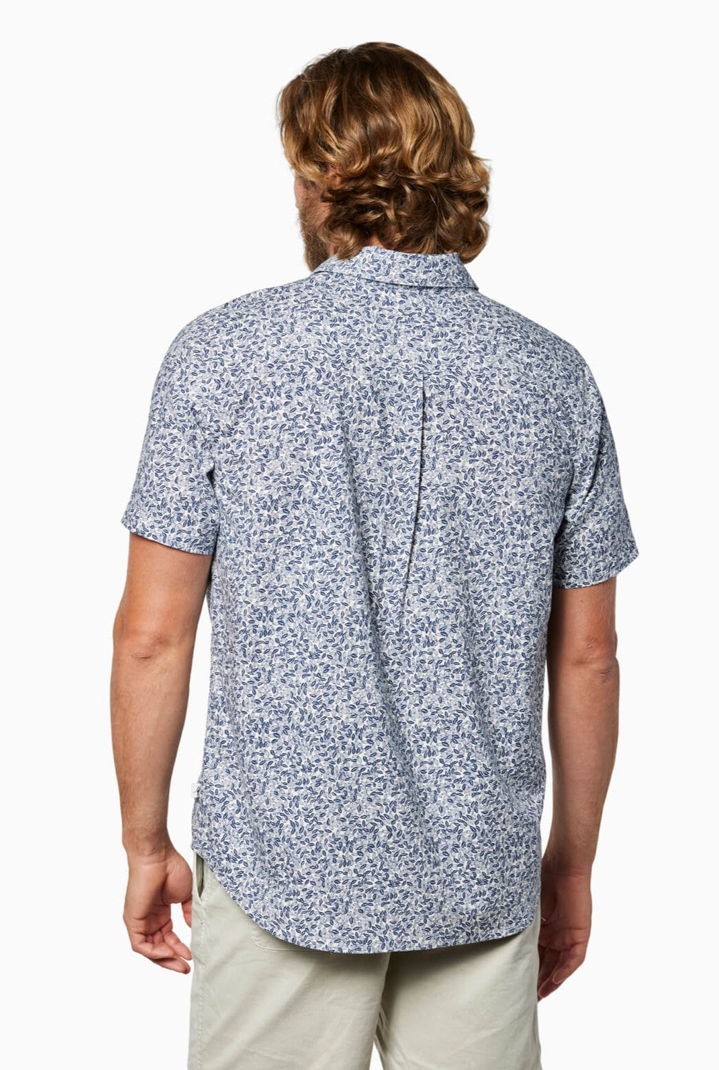 A man with wavy light brown hair is seen from the back in a Toes on the Nose Coastline Short Sleeve Button Up Shirt featuring a light blue and white floral print, paired with light shorts, set against a white background.