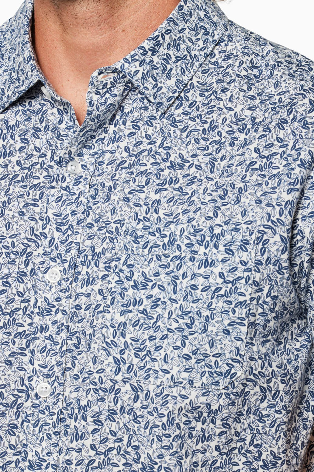 Close-up of a person wearing the Coastline Short Sleeve Button Up Shirt by Toes on the Nose, featuring a blue leafy pattern on a white background and elegant pearl buttons. Only the lower face, neck, and upper chest are shown.