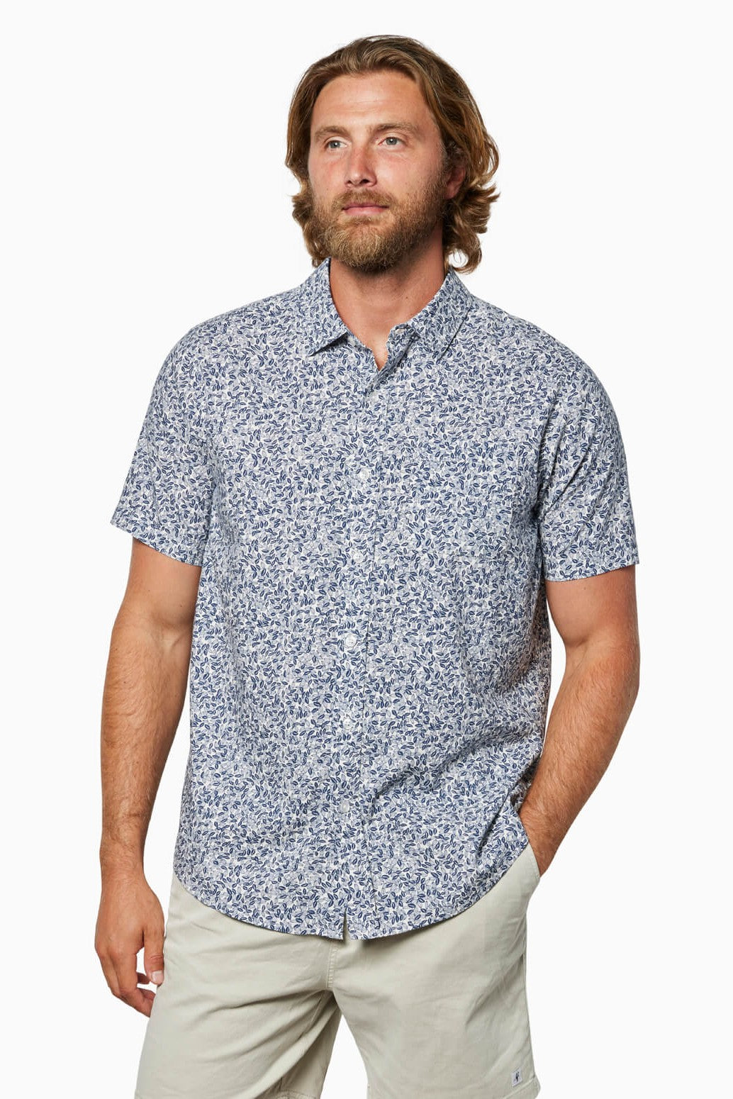 A man with medium-length brown hair and a beard wears the Toes on the Nose Coastline Short Sleeve Button Up Shirt, featuring pearl buttons and a blue and white floral pattern, paired with light shorts, standing against a plain background.