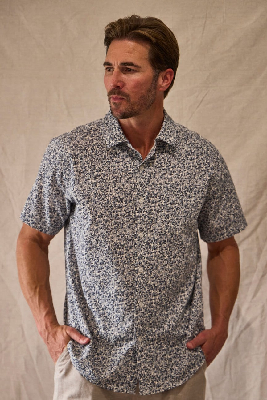 A man with light brown hair and a trimmed beard models the Toes on the Nose Coastline Short Sleeve Button Up Shirt in Navy Multi, featuring pearl buttons, while standing against a beige fabric backdrop with his hands in his pockets.