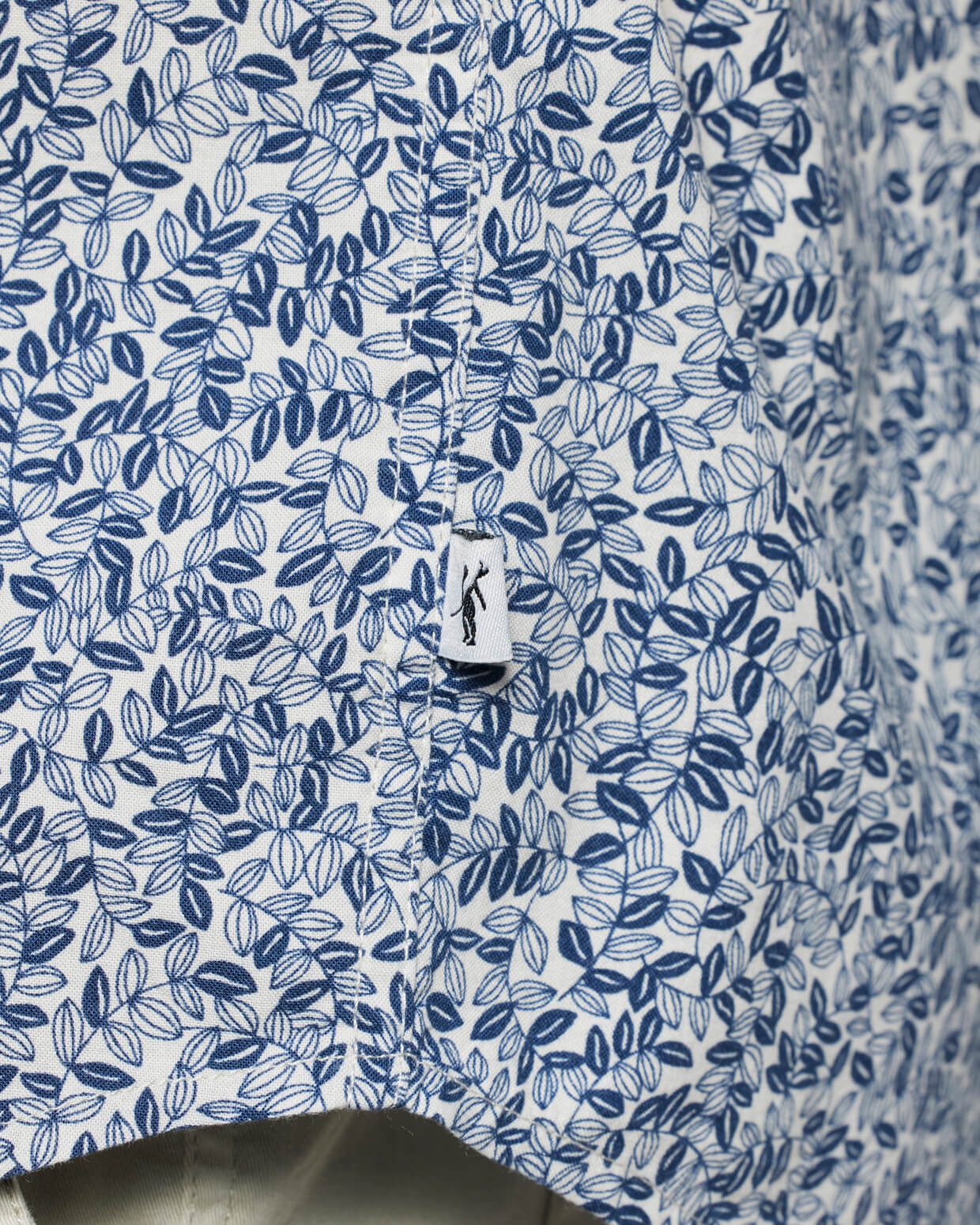 Close-up of the Coastline Short Sleeve Button Up Shirt by Toes on the Nose, featuring a blue and white leafy pattern in soft cotton viscose. The center seam is visible with a small white tag displaying the brand's black logo.