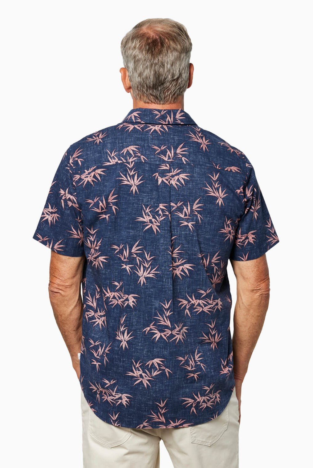 A man with gray hair is shown from behind wearing the Toes on the Nose Coastline Short Sleeve Button Up Shirt, featuring a navy blue base with a pink bamboo pattern and pearl buttons, paired with light shorts against a plain white background.