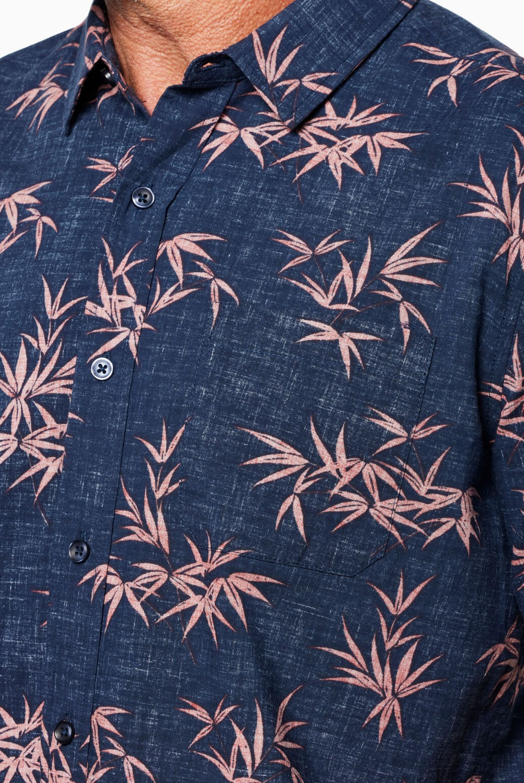 A close-up of a person wearing the Coastline Short Sleeve Button Up Shirt by Toes on the Nose, featuring a navy base with a pink botanical leaf pattern, pearl buttons, textured fabric, and a collar; upper chest and neck are visible.