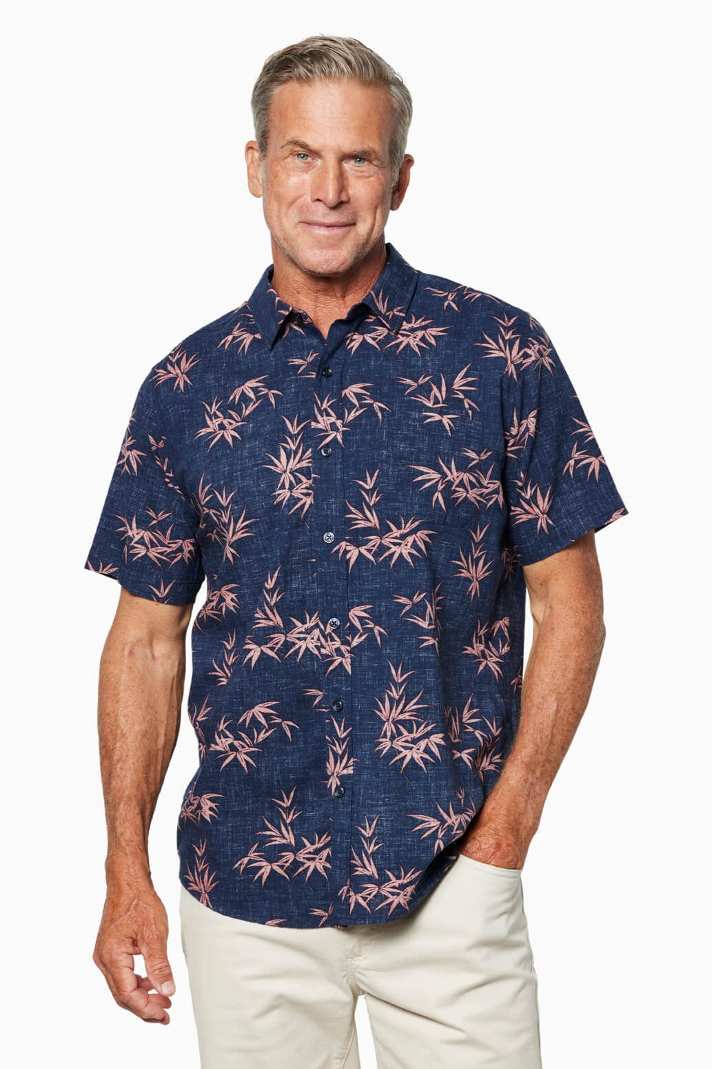 A smiling man with short gray hair wears the Toes on the Nose Coastline Short Sleeve Button Up Shirt in navy with a pink bamboo pattern, pearl buttons, and light-colored pants, standing against a plain white background.