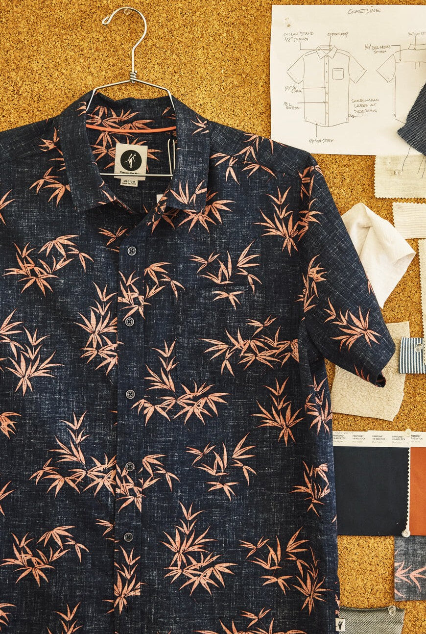 The Coastline Short Sleeve Button Up Shirt by Toes on the Nose, featuring a navy blue and pink bamboo leaf pattern with pearl buttons, hangs on a corkboard amid fabric swatches and hand-drawn design sketches.