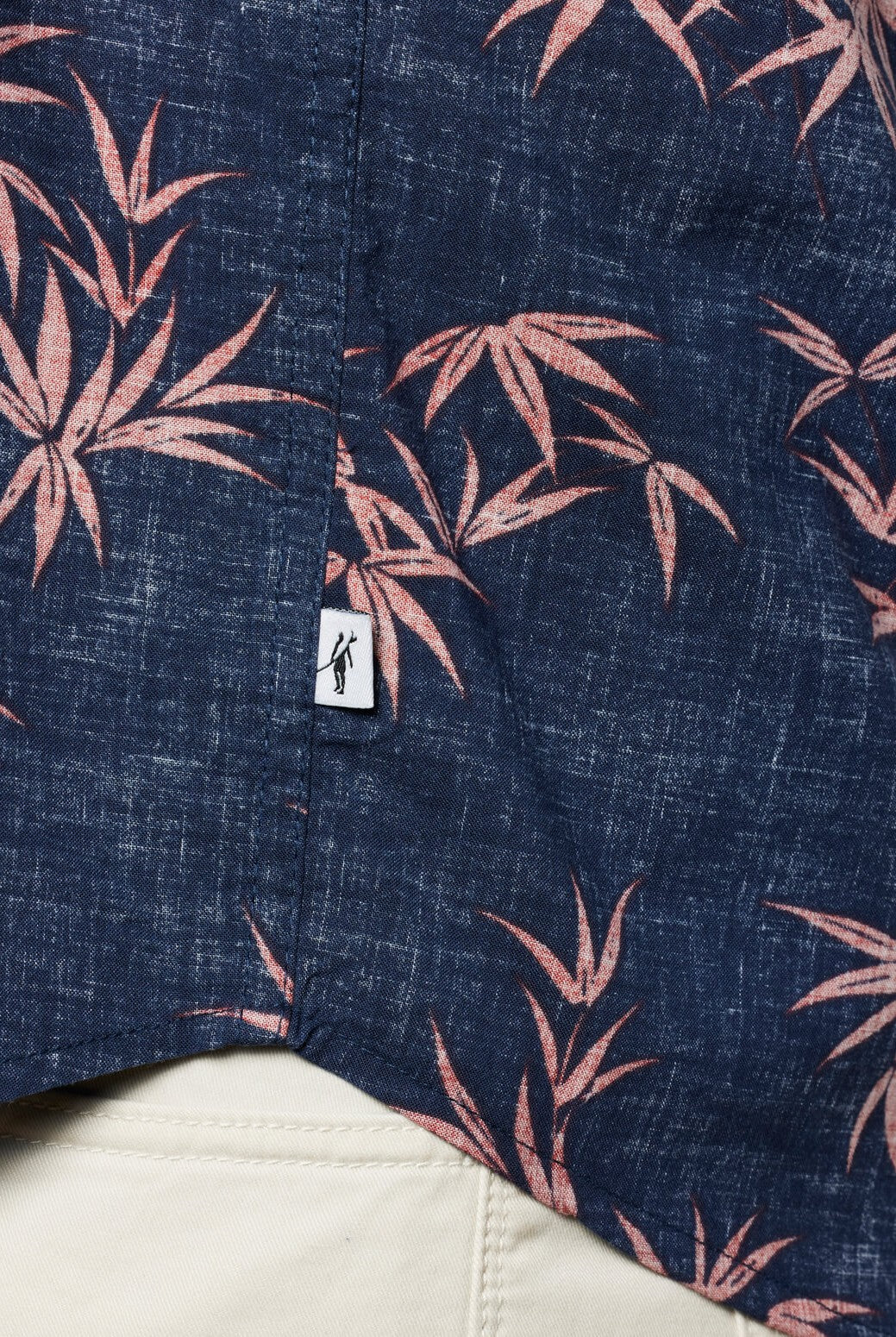 A close-up of the Toes on the Nose Coastline Short Sleeve Button Up Shirt in navy with a pink tropical leaf pattern, pearl buttons, and a small white tag, styled over beige pants.