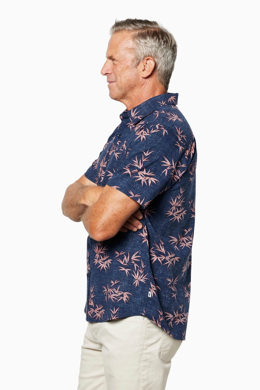 A middle-aged man in profile, arms crossed, wears the Toes on the Nose Coastline Short Sleeve Button Up Shirt with a pink tropical leaf pattern, pearl buttons, and chest pocket, paired with light pants against a plain white background.