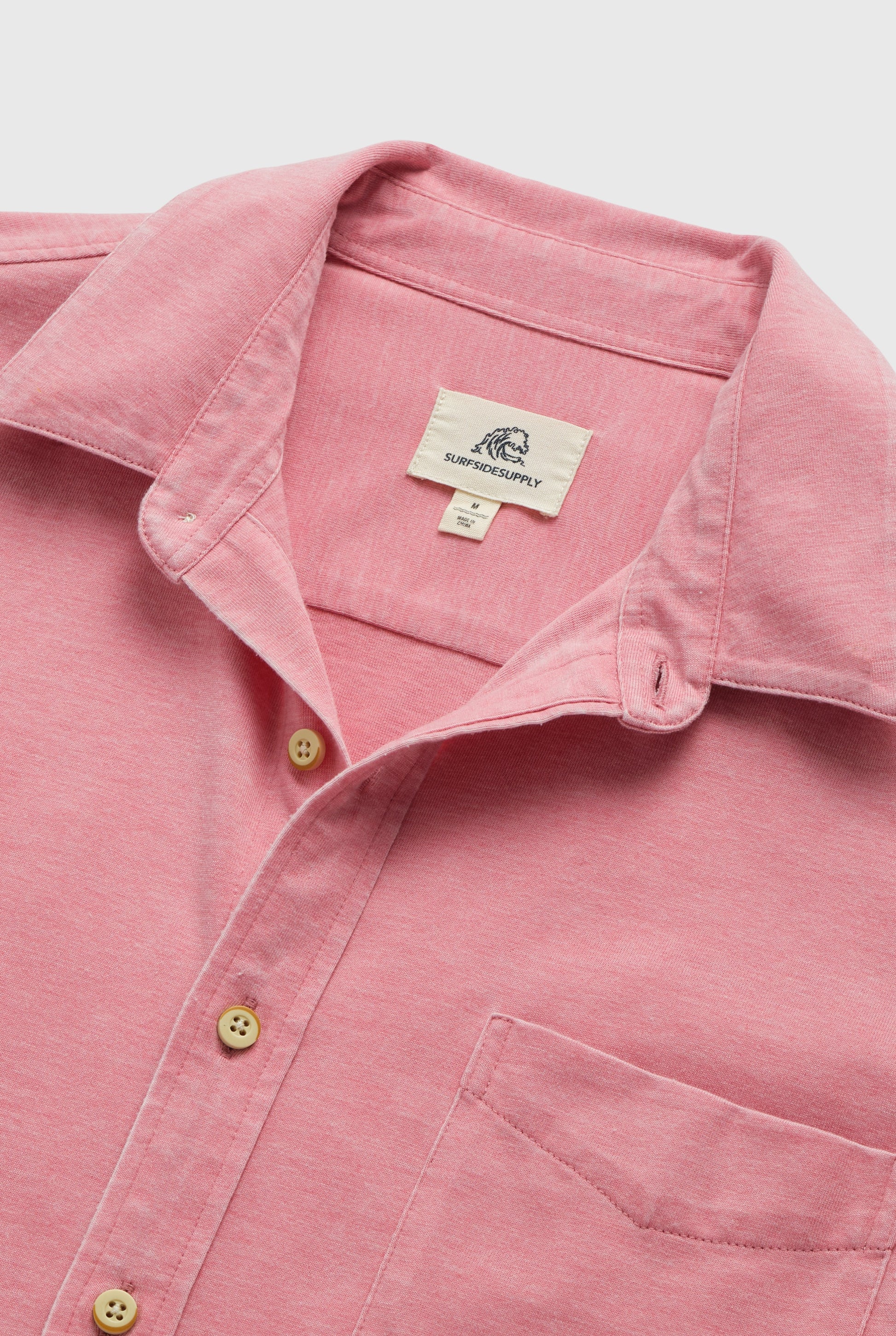A close-up of the Tony Short Sleeve Burnout Knit Shirt in pink by Surfside Supply Co., featuring a front pocket, beige buttons, and a regular fit. The collar label shows the brand name with a wave logo above it.
