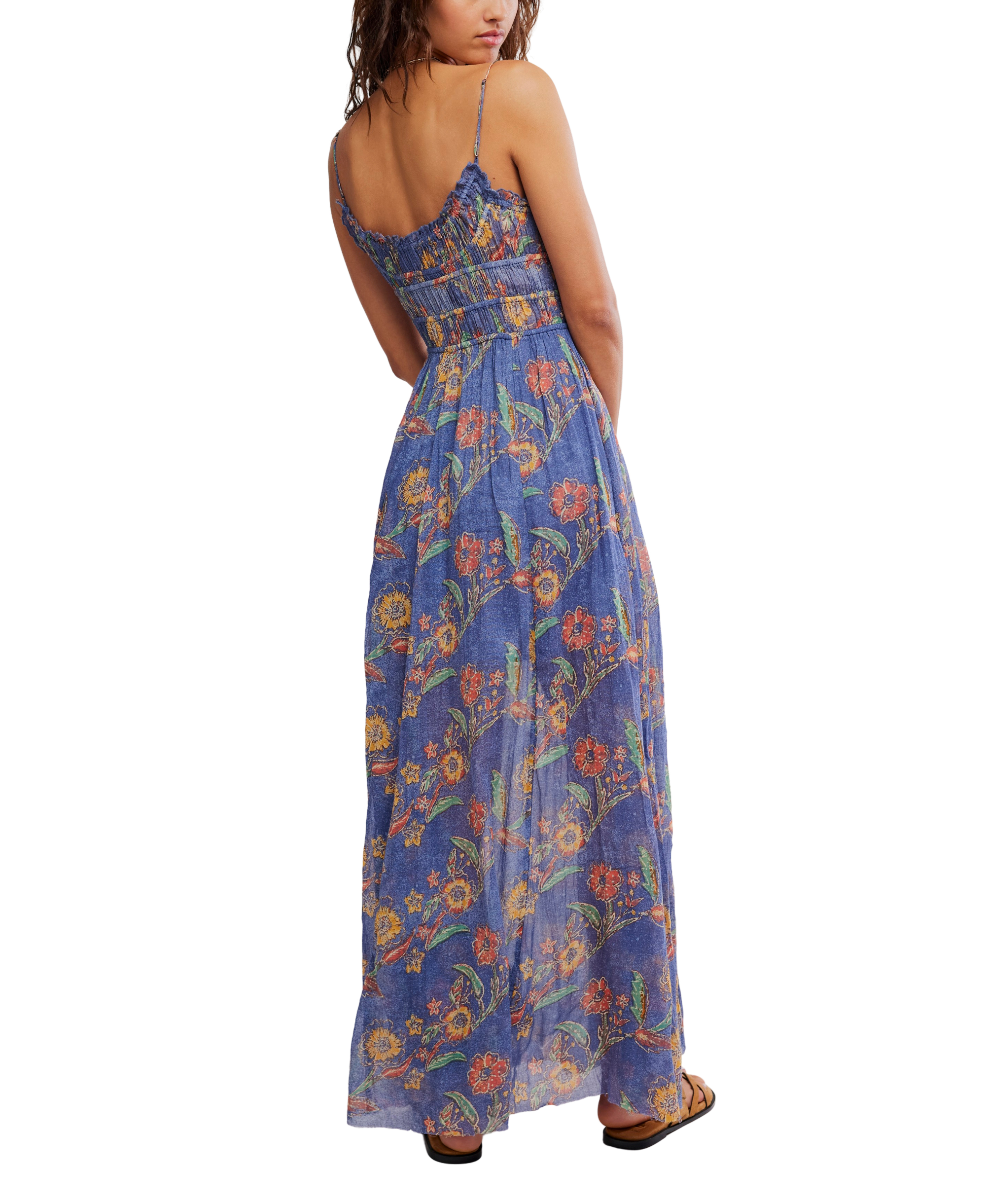 A woman with wavy hair stands facing away in the Free People Fp One Ciella Printed Maxi—a sleeveless, blue floral dress with adjustable straps. She wears sandals, and the background is white.