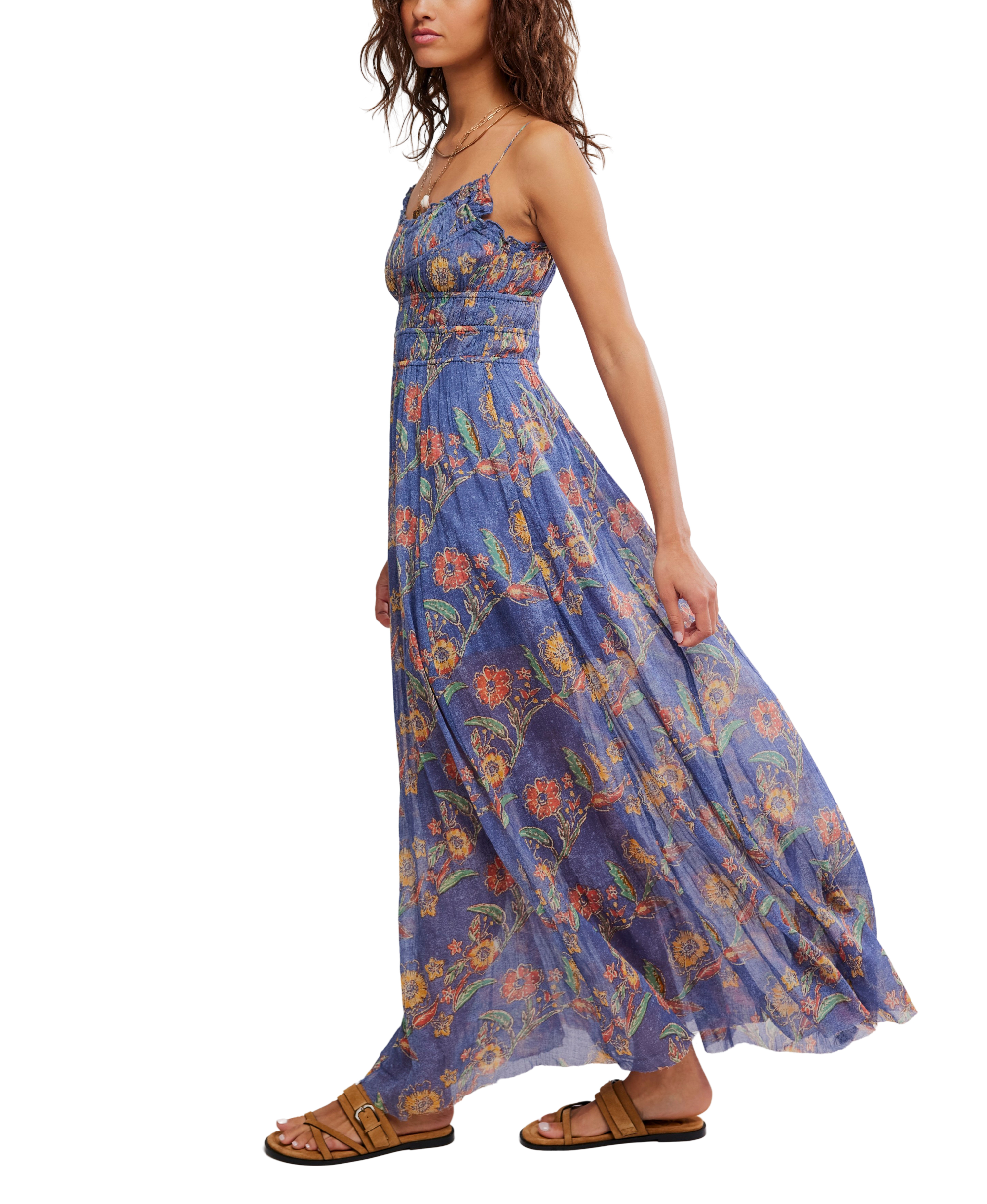 A woman in the Free People Fp One Ciella Printed Maxi, a sleeveless blue cotton dress with a scoop neck and colorful floral print, walks in brown sandals while holding part of her flowing skirt.