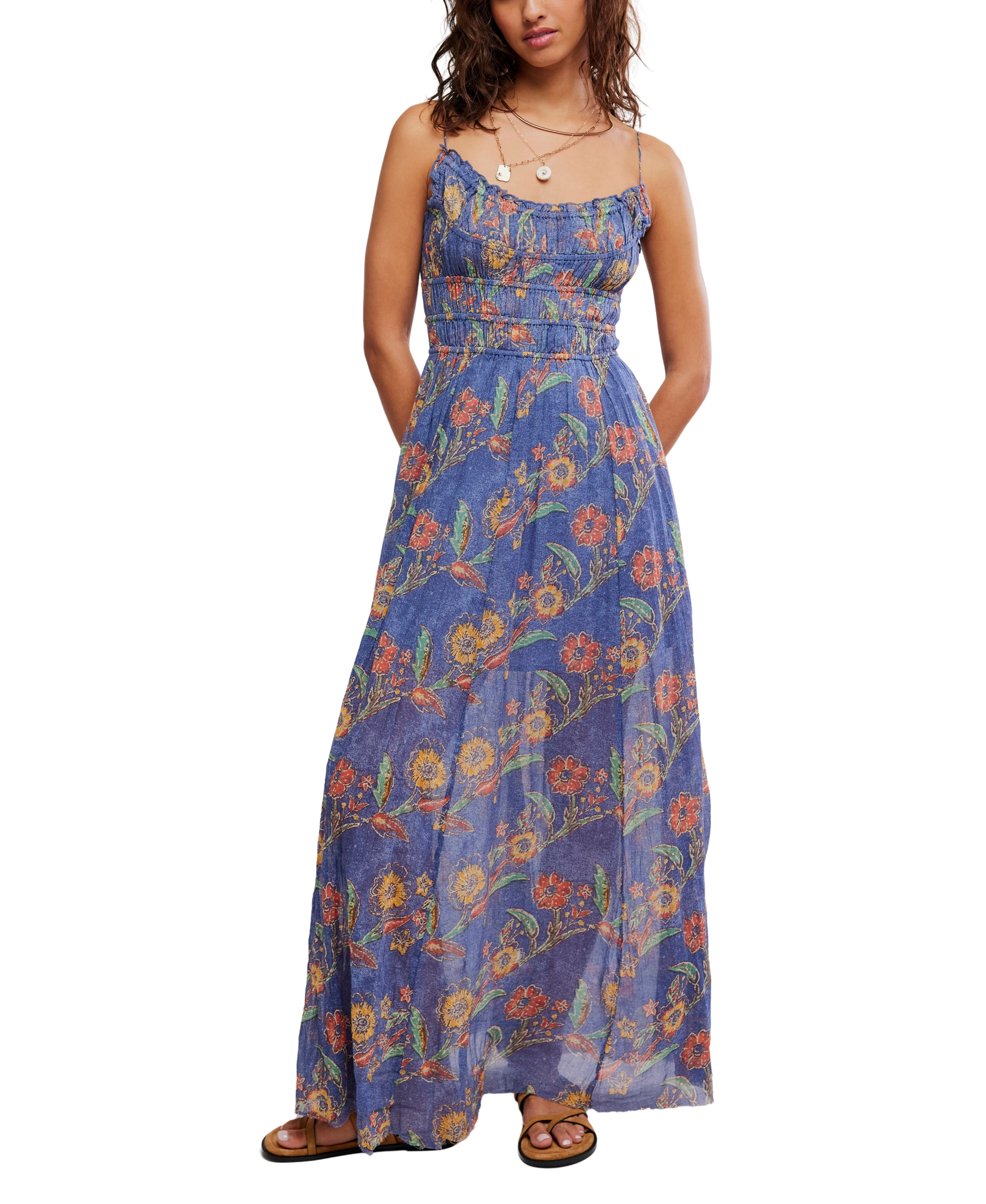 A woman stands with her hands behind her back wearing the Free People Fp One Ciella Printed Maxi in Blue Multi, featuring a sleeveless scoop neck, adjustable straps, colorful floral pattern, sandals, and layered necklaces.
