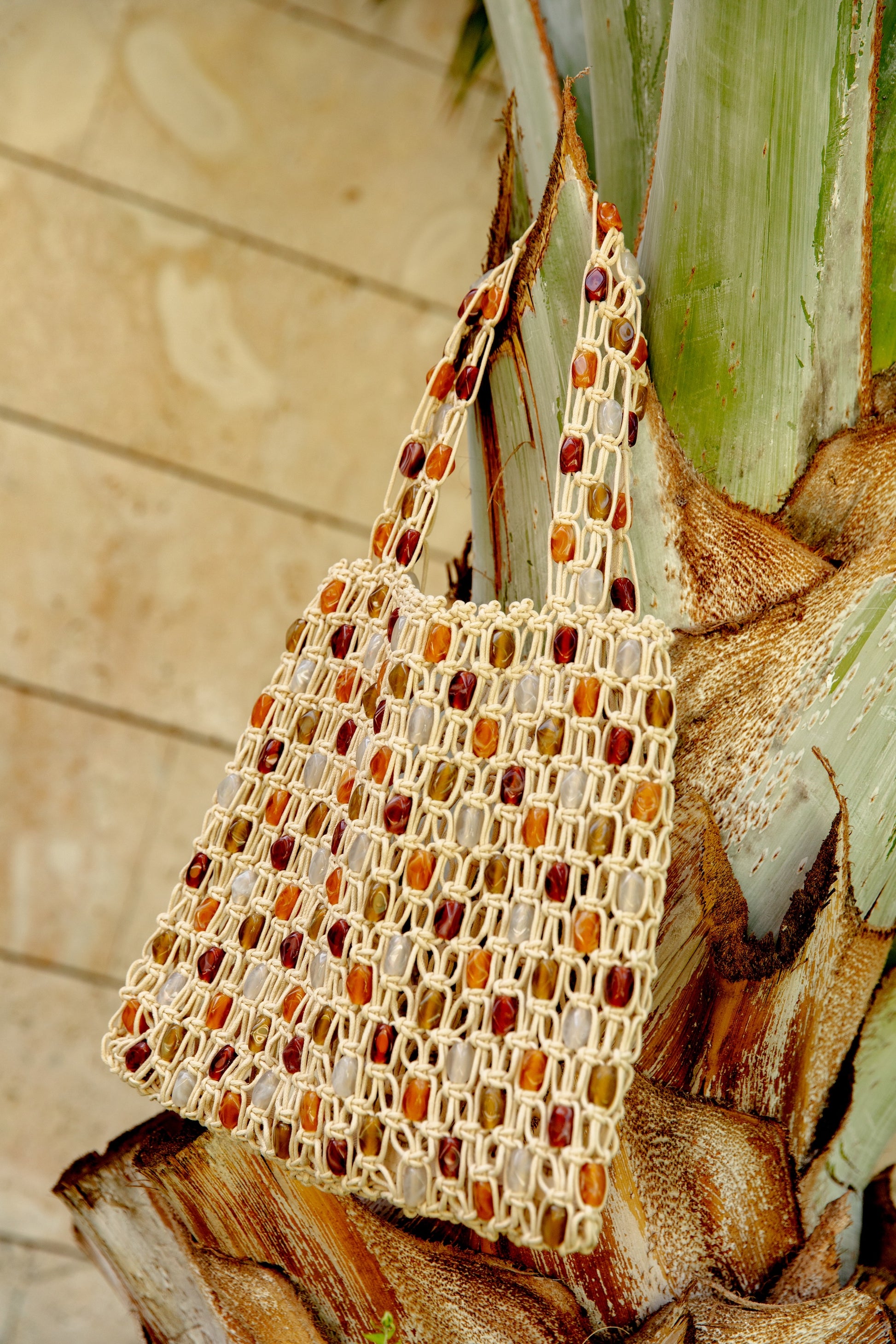 The Asher Beaded Beach Bag by Urban Expressions, featuring handcrafted boho style and wooden beads in natural and brown tones, hangs on a palm tree trunk with a blurred stone wall behind it.
