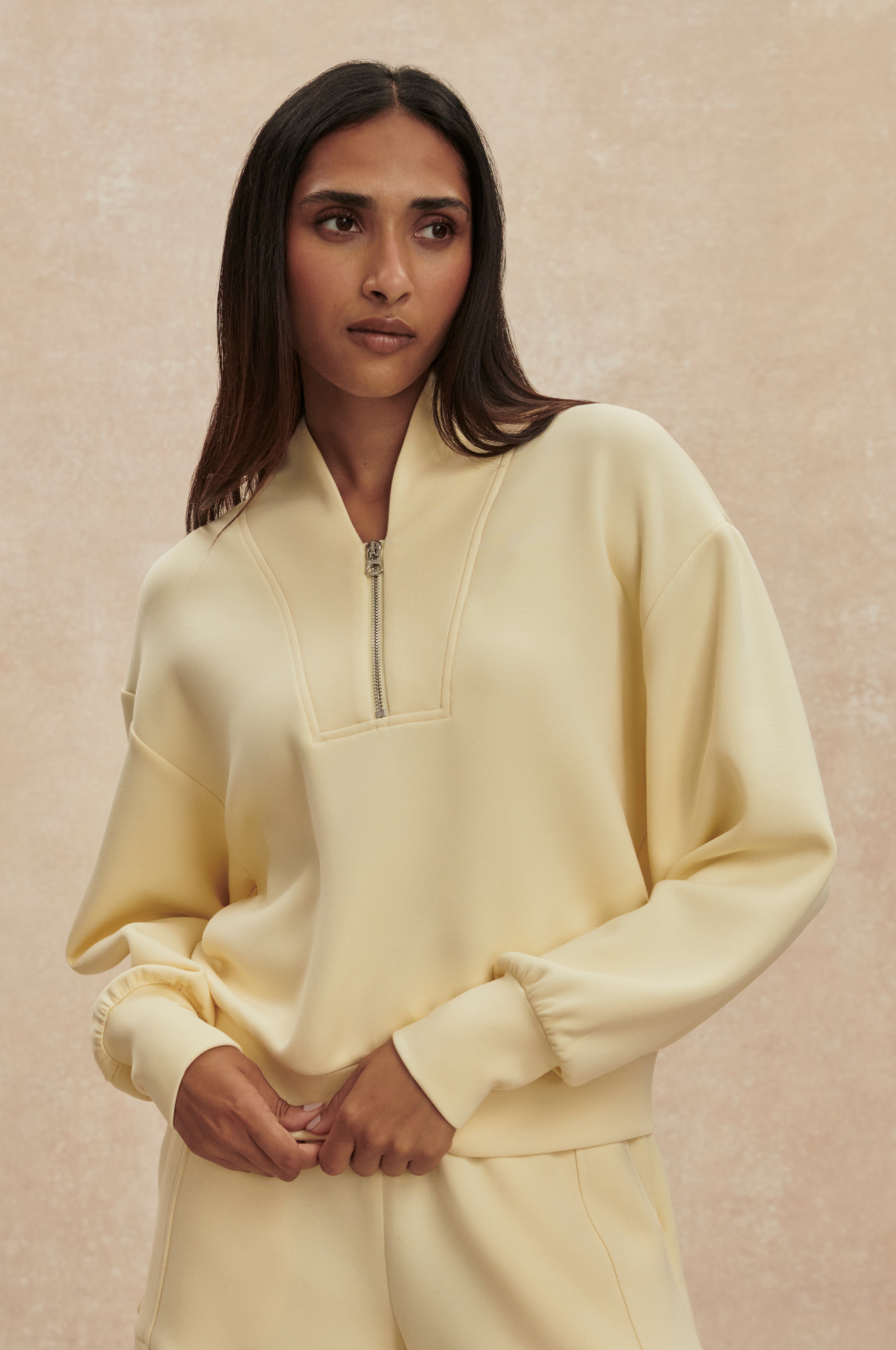 A woman with long dark hair wears the Varley Davidson Sweat Top, a light yellow zip-up pullover with a standing collar, paired with matching pants. She stands against a neutral beige background, looking calm.
