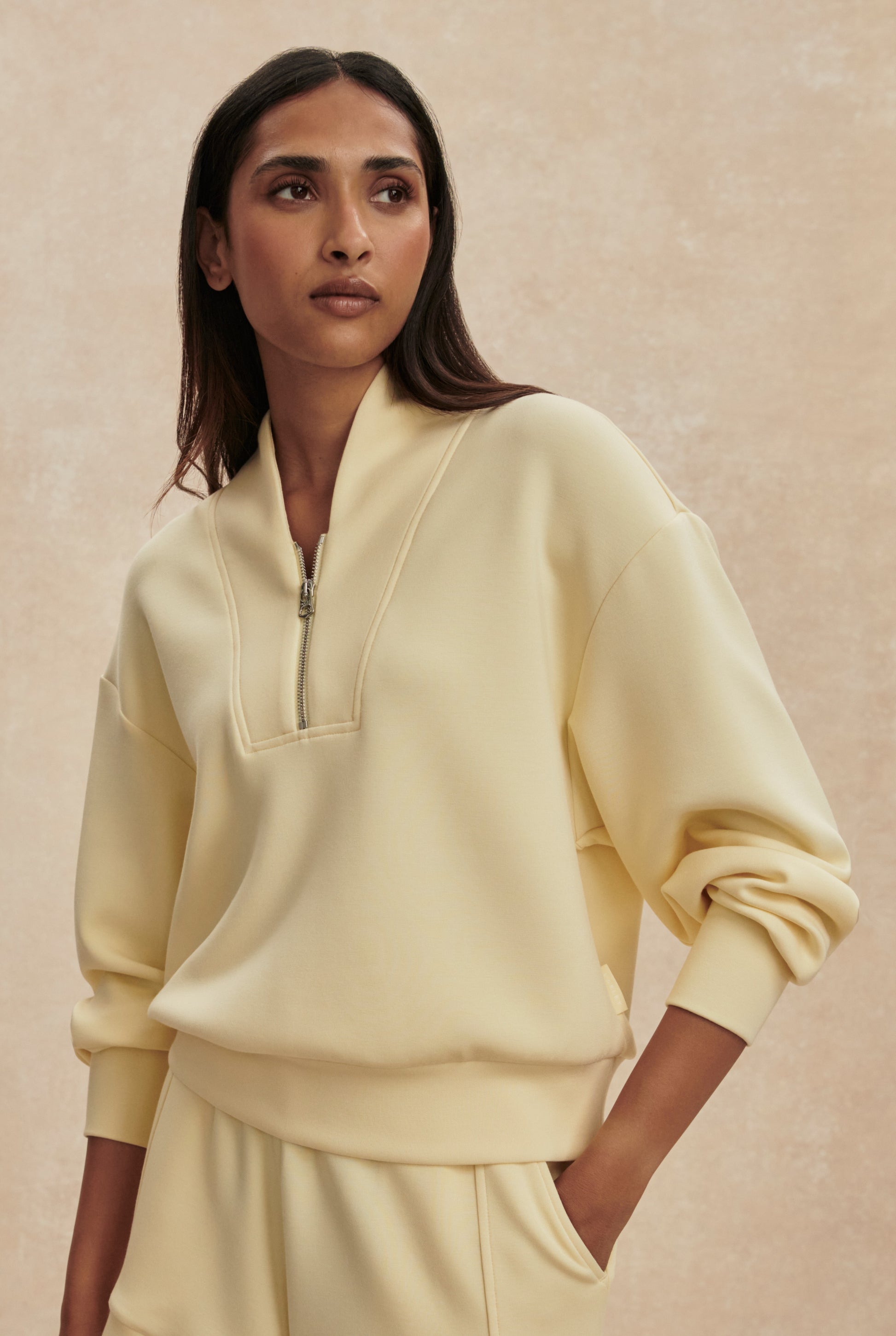 A woman with long dark hair wears the Varley Davidson Sweat Top, a pale yellow relaxed-fit zip-up pullover with a standing collar, paired with matching pants, standing against a beige background and looking calmly to the left.
