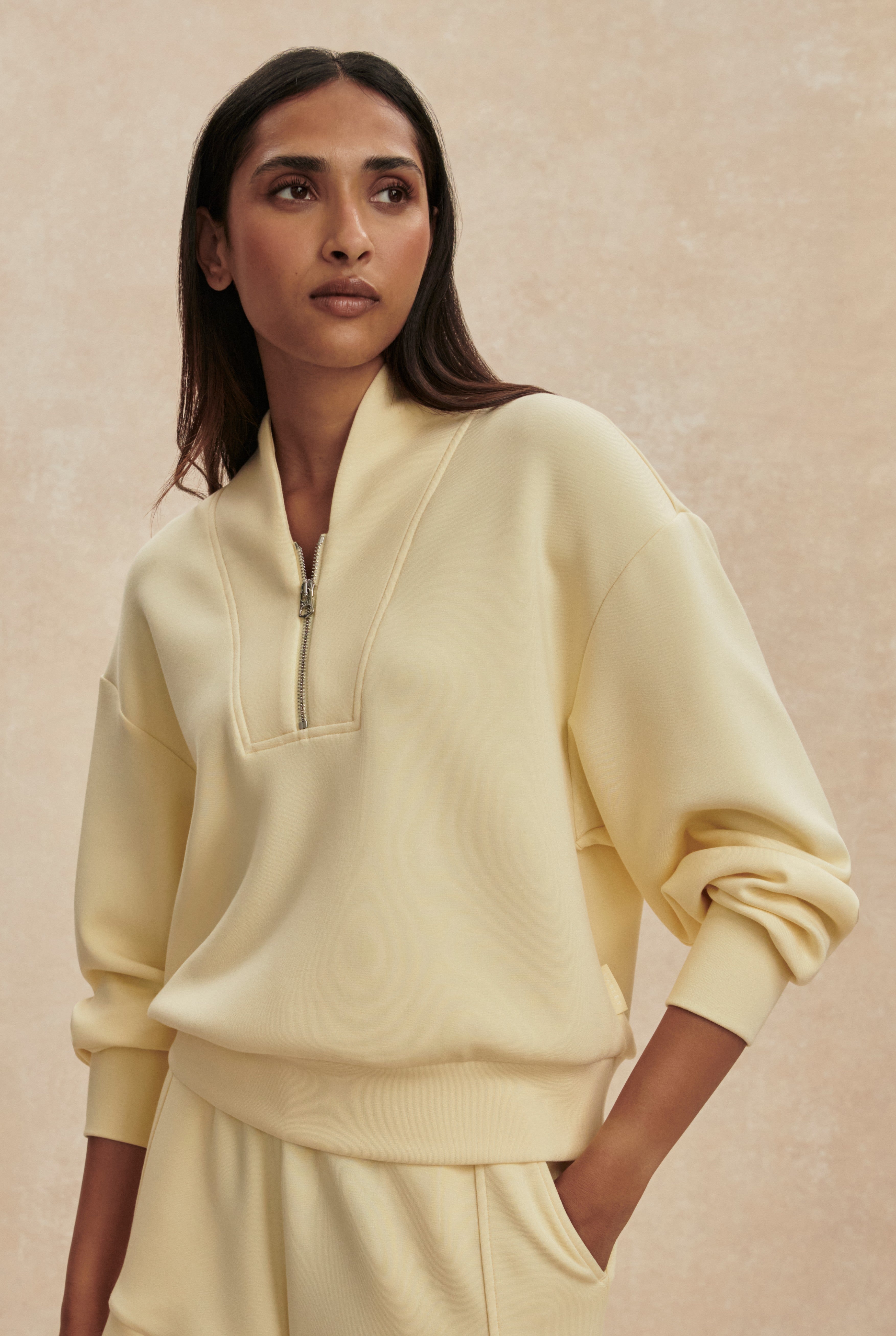 A woman with long dark hair wears the Varley Davidson Sweat Top, a pale yellow relaxed-fit zip-up pullover with a standing collar, paired with matching pants, standing against a beige background and looking calmly to the left.
