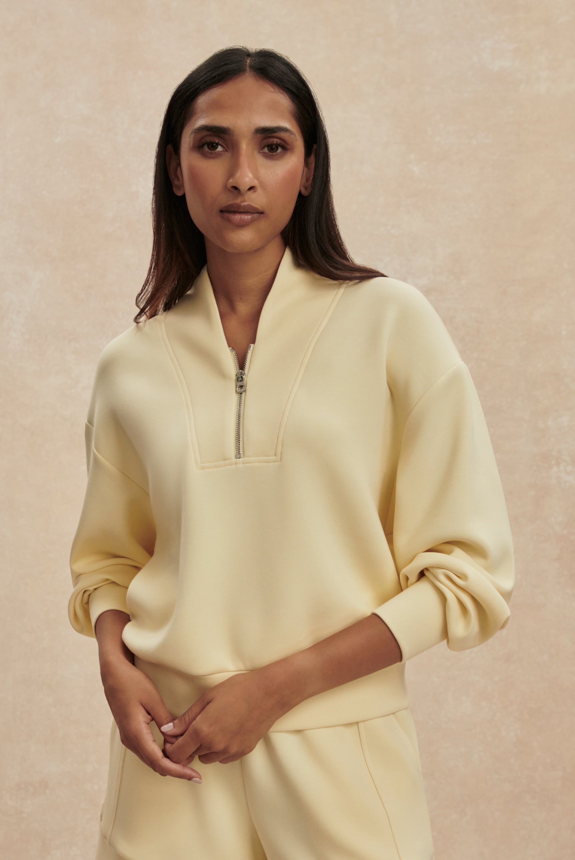 Against a light beige background, a person with long dark hair and a calm expression wears Varley's Davidson Sweat Top in yellow, paired with matching pants. One hand rests on their wrist.