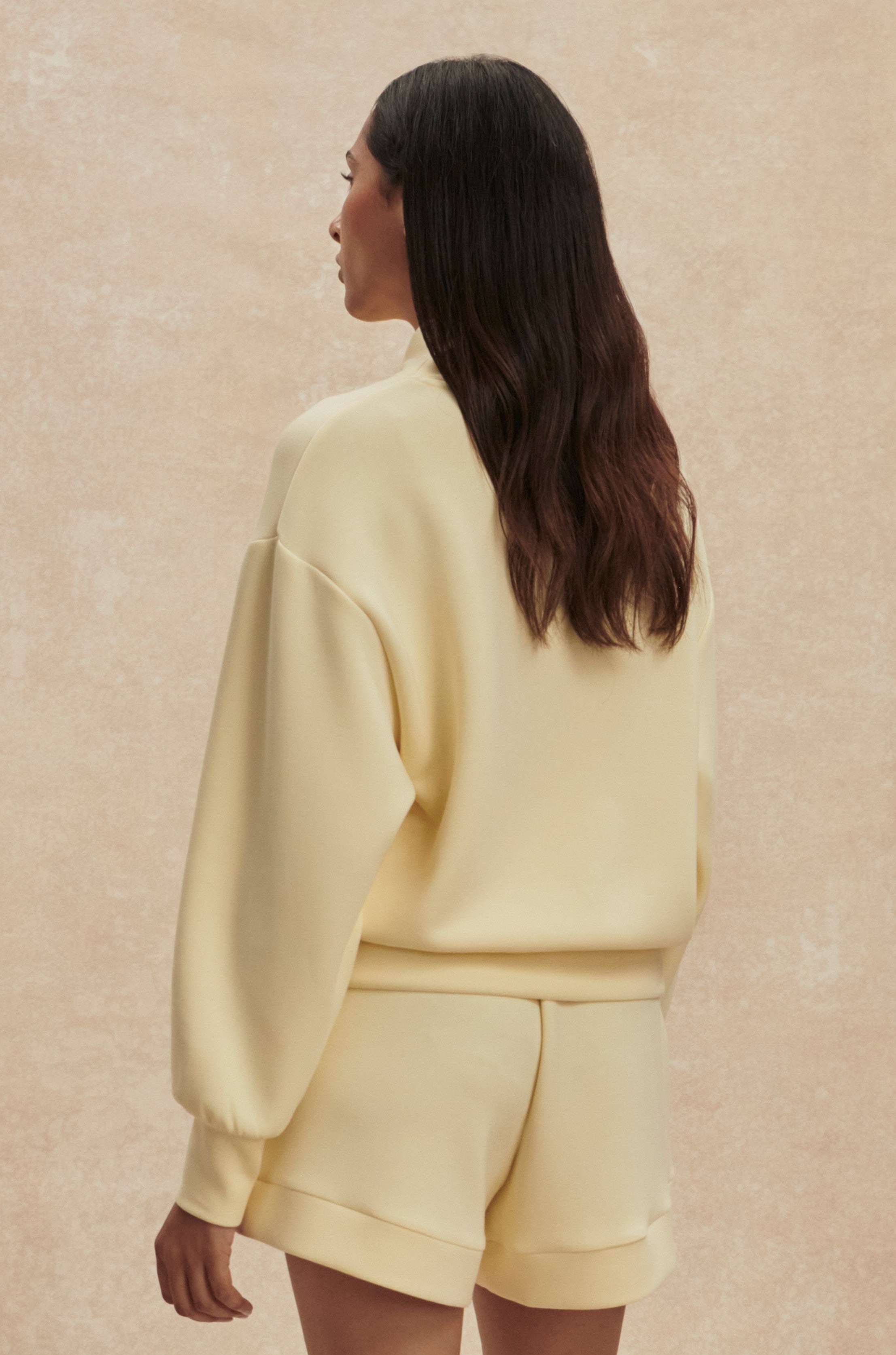 A woman with long, wavy dark hair stands facing away, wearing the Varley Davidson Sweat Top in pale yellow and matching shorts against a light beige textured background.