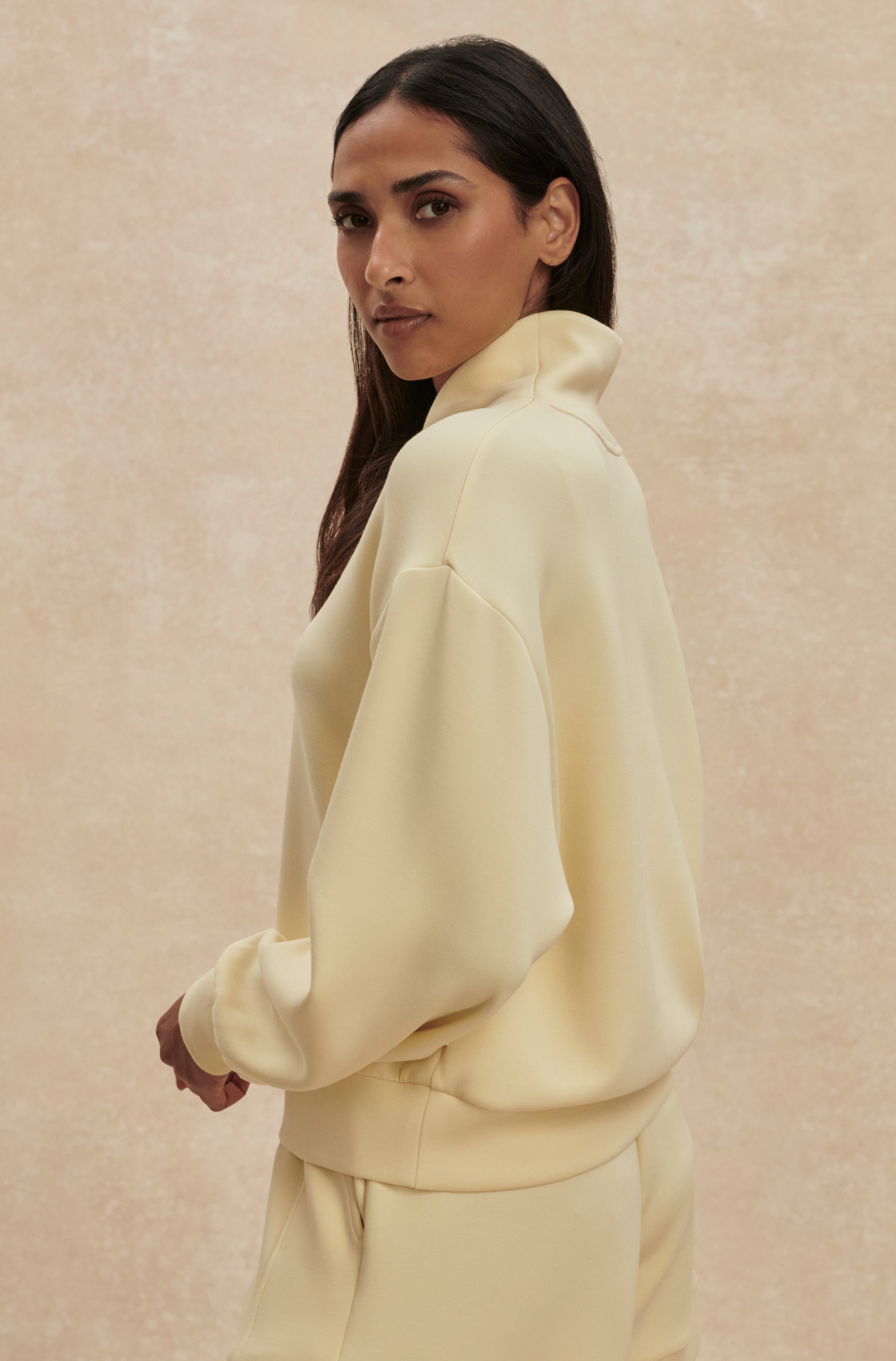 A woman with long dark hair, wearing the Varley Davidson Sweat Top and matching relaxed-fit pants in light yellow, stands sideways and looks over her shoulder against a beige textured background.