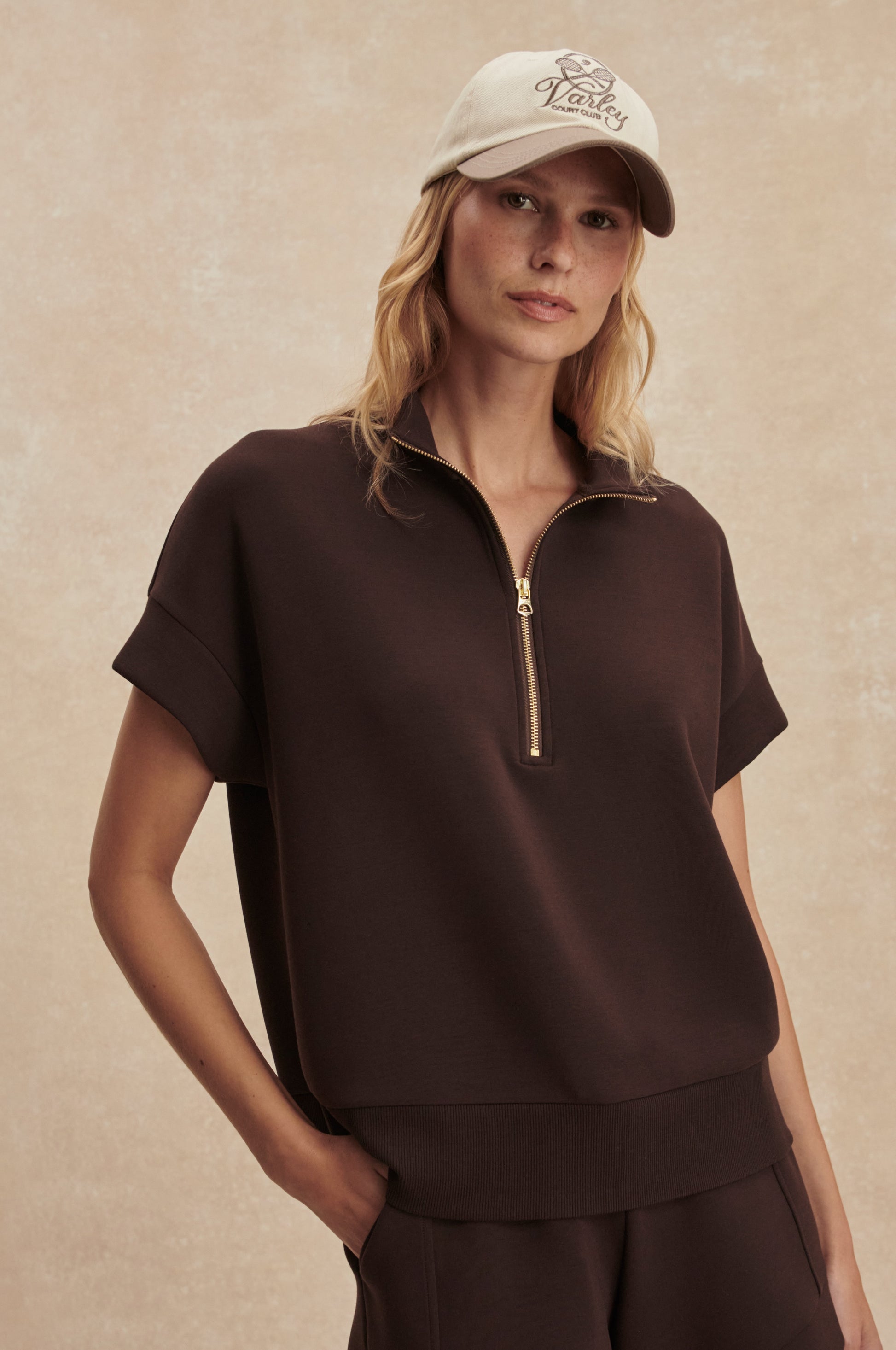 A woman in the Varley Ritchie Short Sleeve Sweat in brown with matching pants and a beige cap, standing against a neutral textured background.