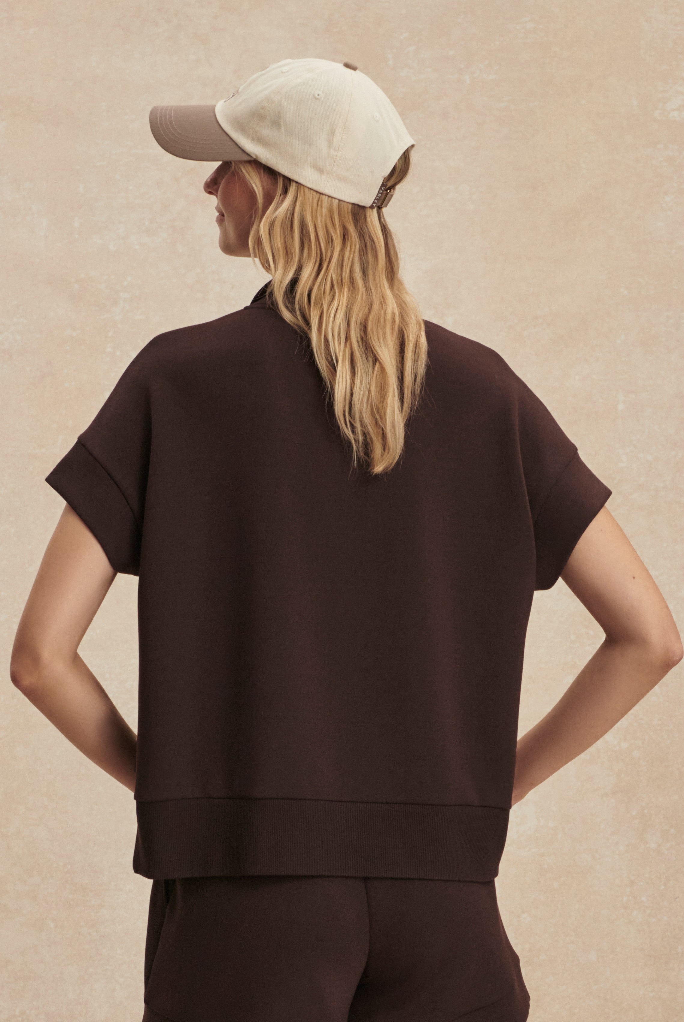 A person with long, wavy blonde hair wears the Varley Ritchie Short Sleeve Sweat in dark brown and matching pants, plus a beige baseball cap, shown from the back against a neutral background.
