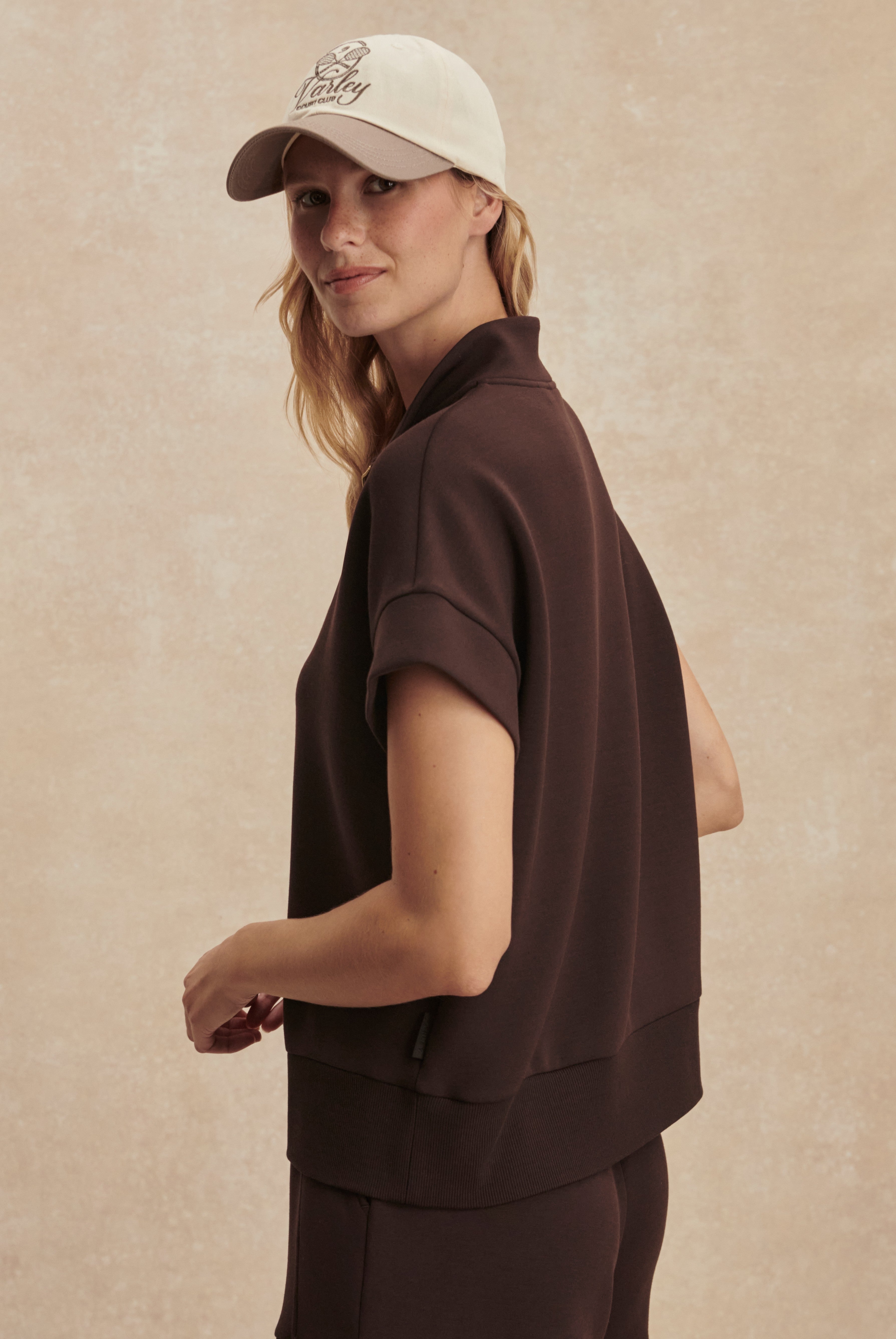 A woman wears the Varley Ritchie Short Sleeve Sweat in dark brown with matching pants and a light-colored baseball cap, standing sideways and smiling slightly against a beige textured background.