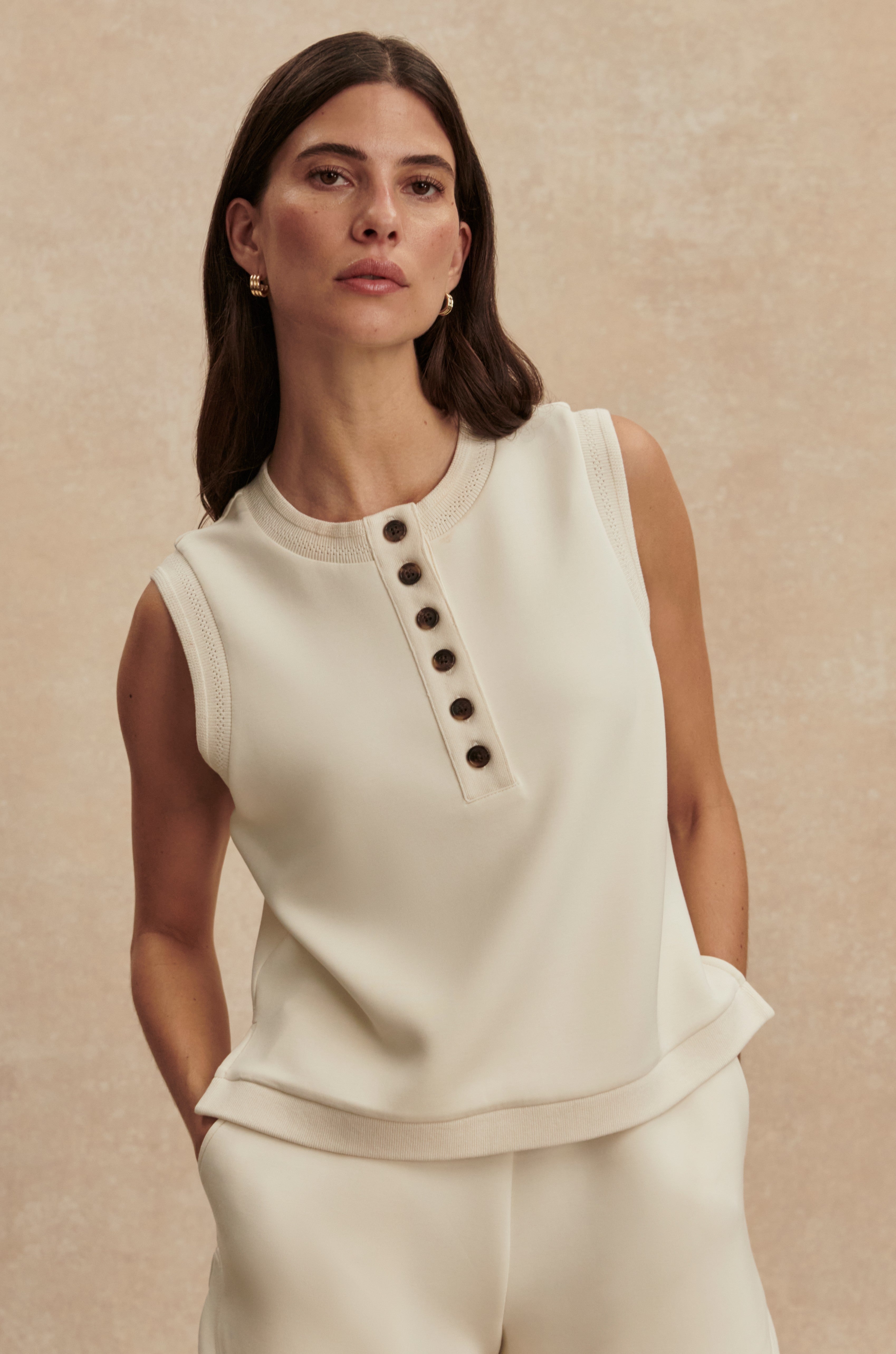A woman with long brown hair wears the Varley Marla Button Placket Tank in cream DoubleSoft fabric with decorative trim, standing against a neutral textured background. She has a neutral expression and small hoop earrings.