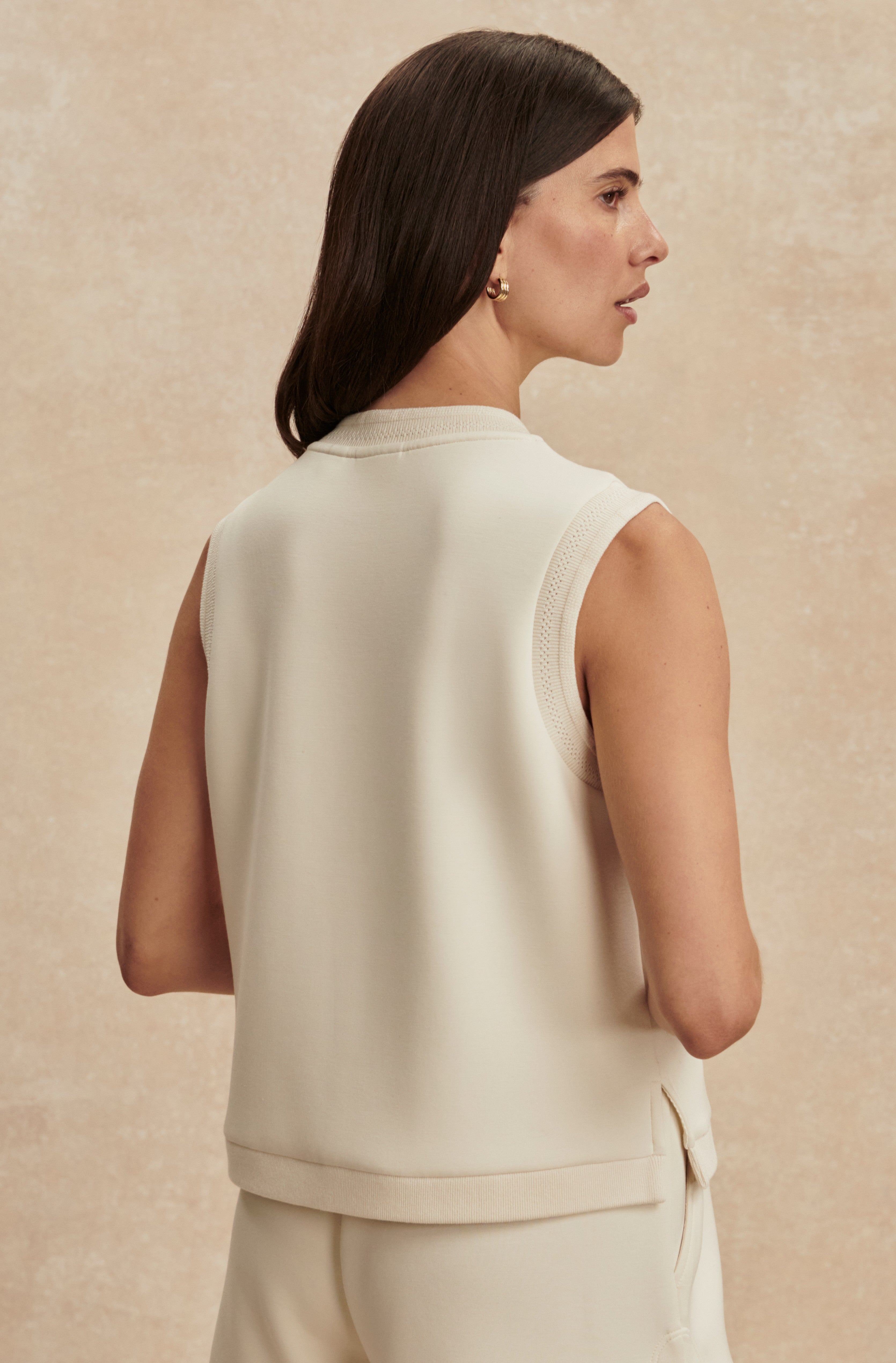 A woman with straight brown hair, wearing the Varley Marla Button Placket Tank and matching DoubleSoft pants, stands with her back to the camera against a beige background.