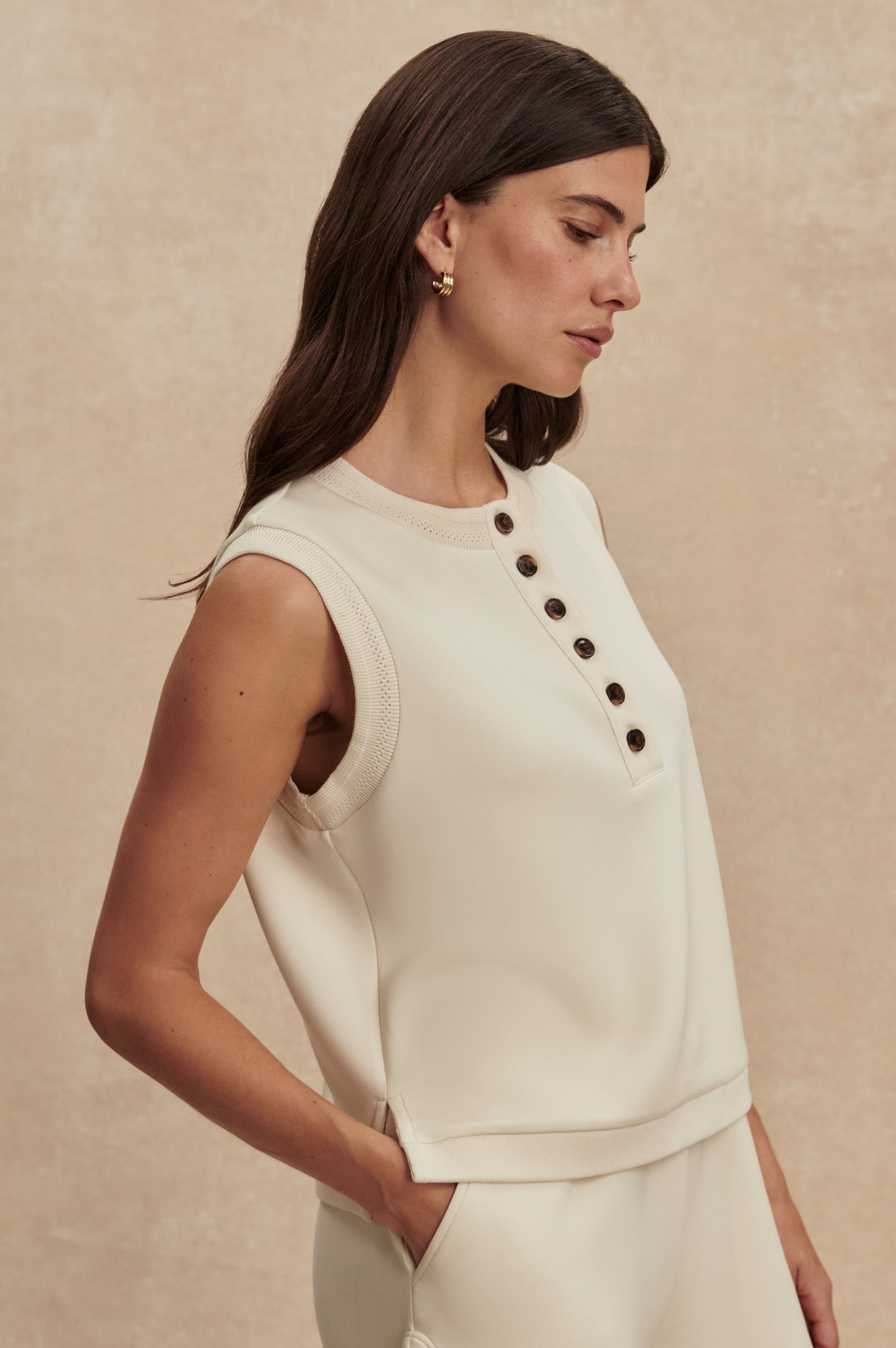 A woman with long brown hair wears the Varley Marla Button Placket Tank in DoubleSoft fabric with black buttons and matching pants, posing in profile against a beige background.
