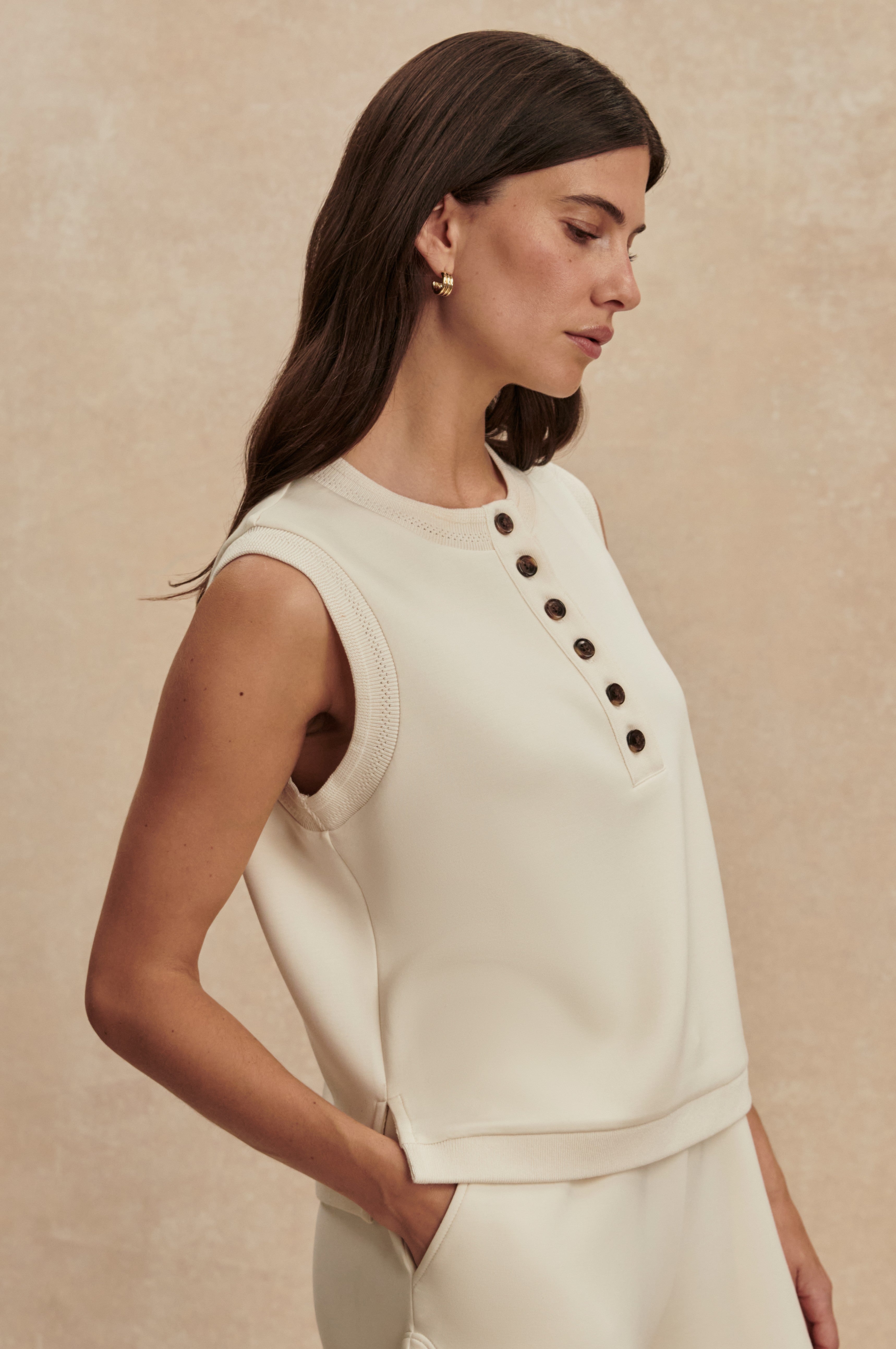A woman with long brown hair wears the Varley Marla Button Placket Tank in DoubleSoft fabric with black buttons and matching pants, posing in profile against a beige background.
