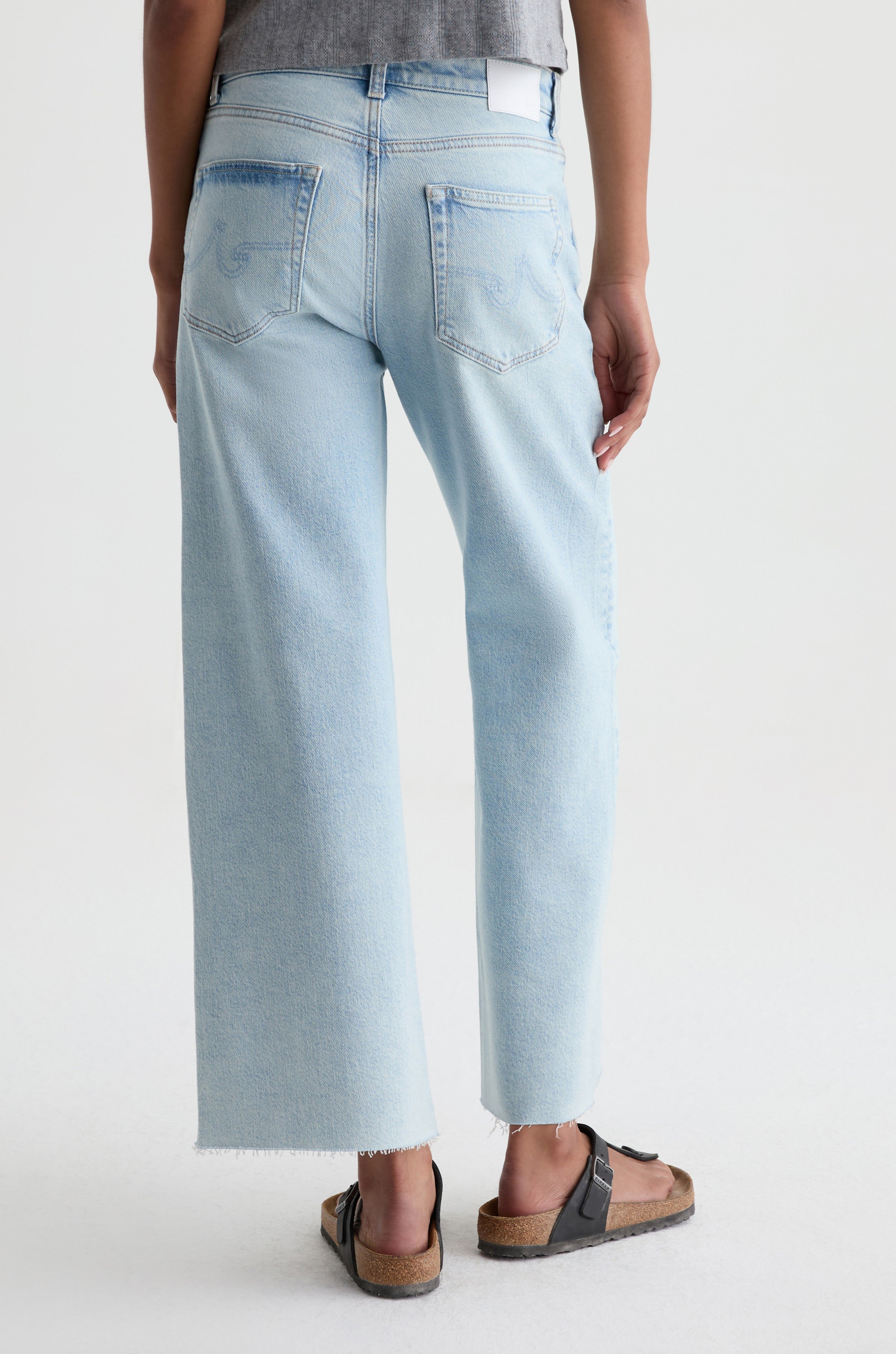 A person faces away, wearing AG Jeans Saige Crop Wide Leg in Harvest—light-wash, relaxed jeans with raw hems and cropped legs—paired with a gray top and brown sandals, set against a plain light background.