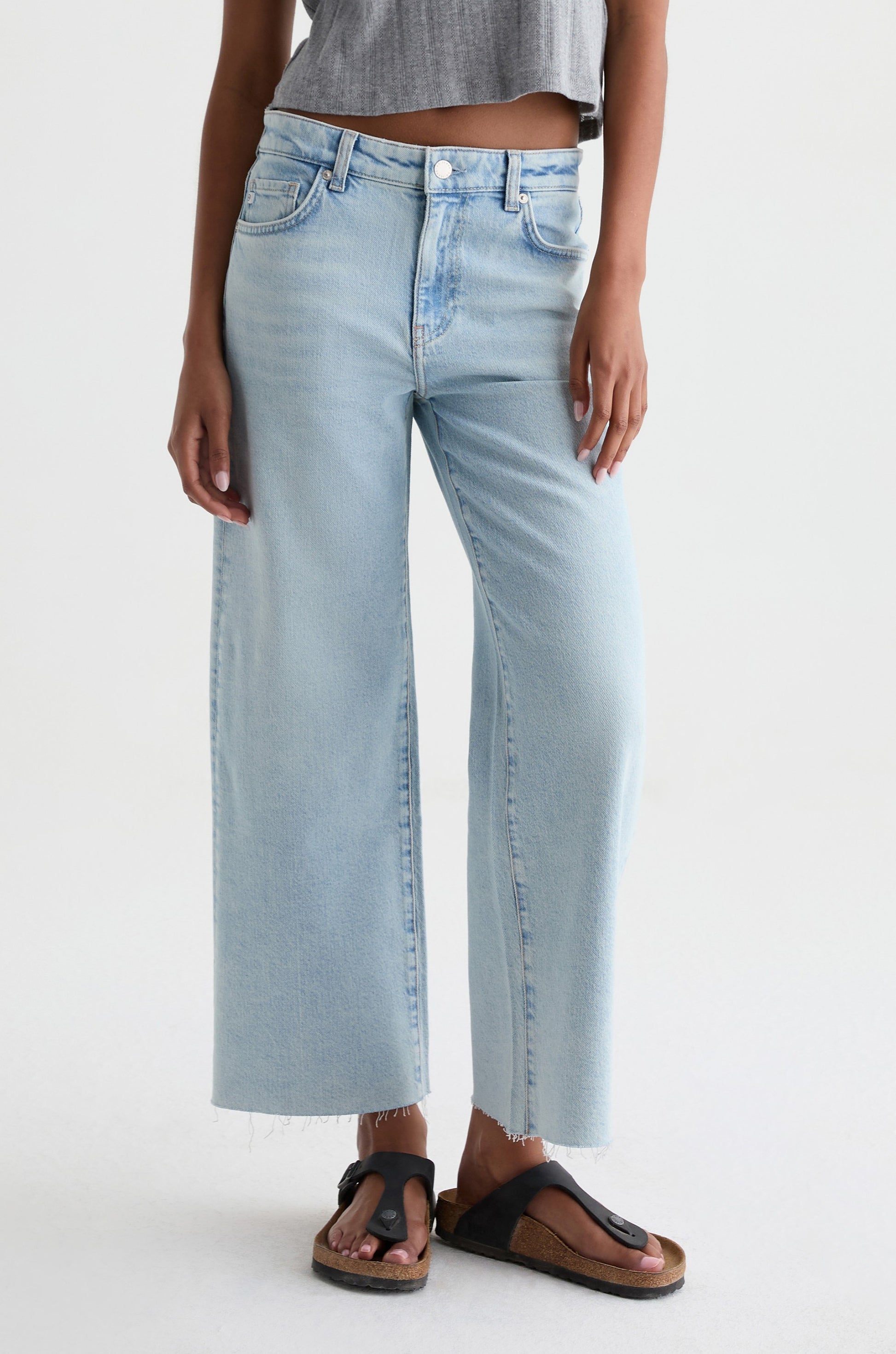 A person wears AG Jeans' Saige Crop Wide Leg jeans in HARVEST, styled with a gray crop top and black sandals with cork soles, standing against a plain white background.