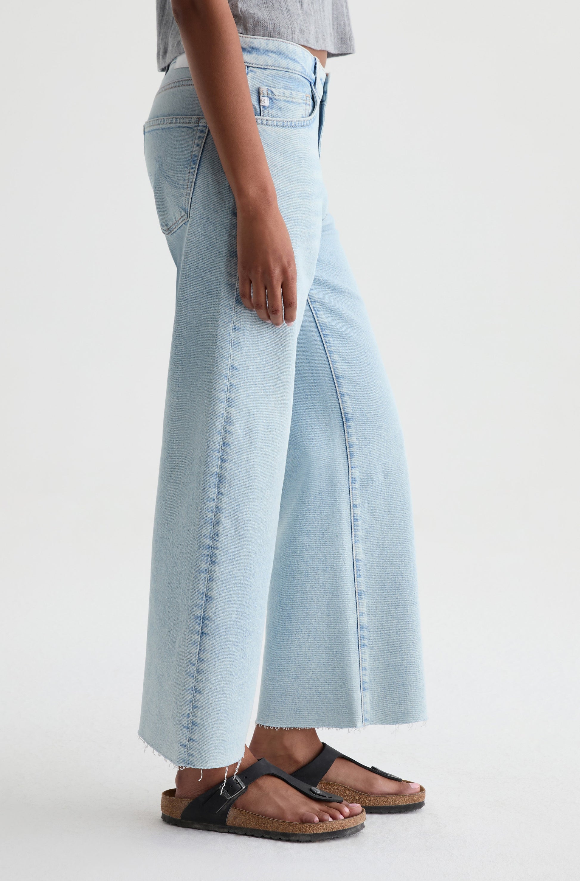 A person wearing AG Jeans’ Saige Crop Wide Leg in Harvest, a grey top, and black sandals stands against a plain white background.