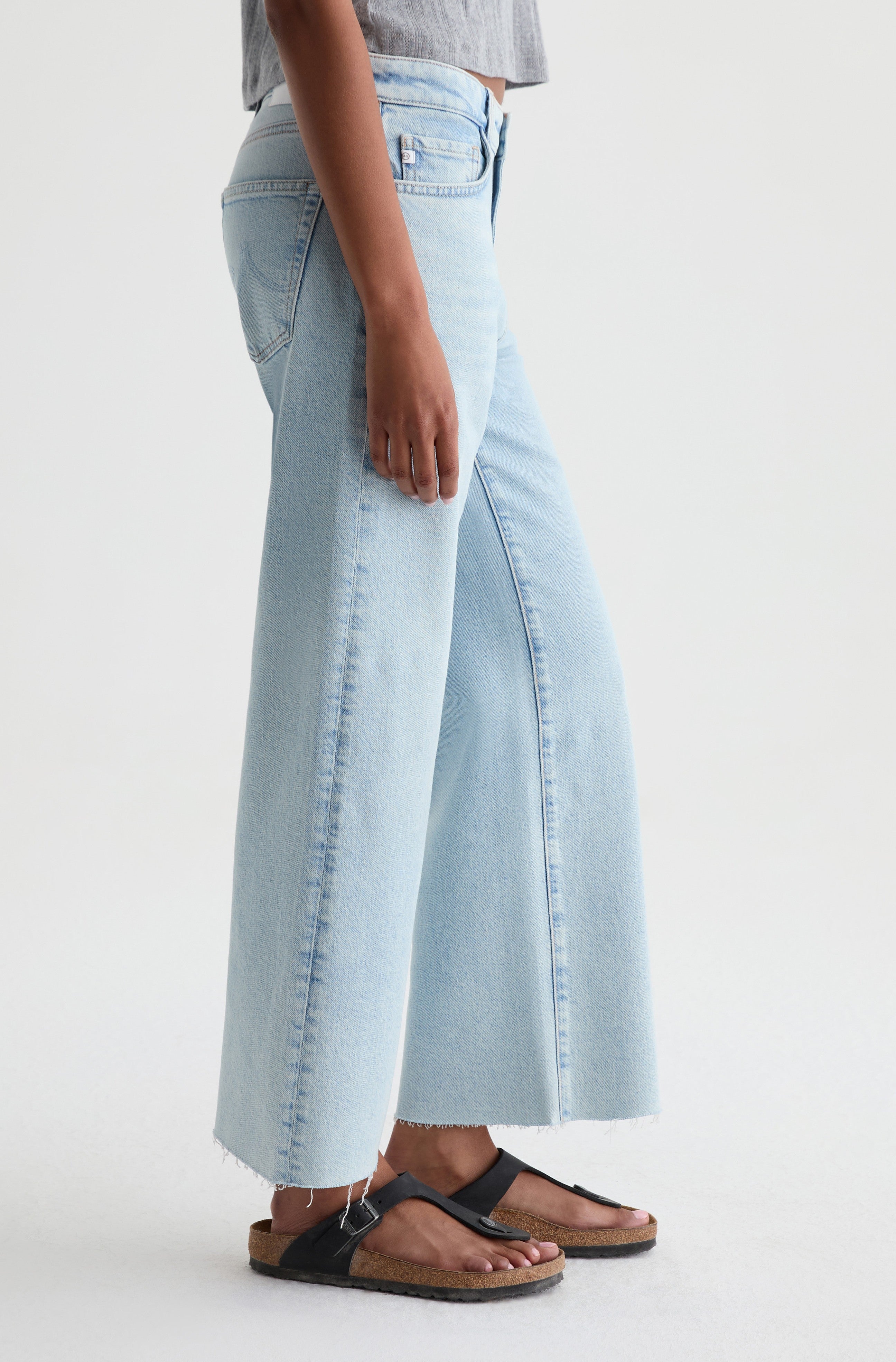 A person wearing AG Jeans’ Saige Crop Wide Leg in Harvest, a grey top, and black sandals stands against a plain white background.