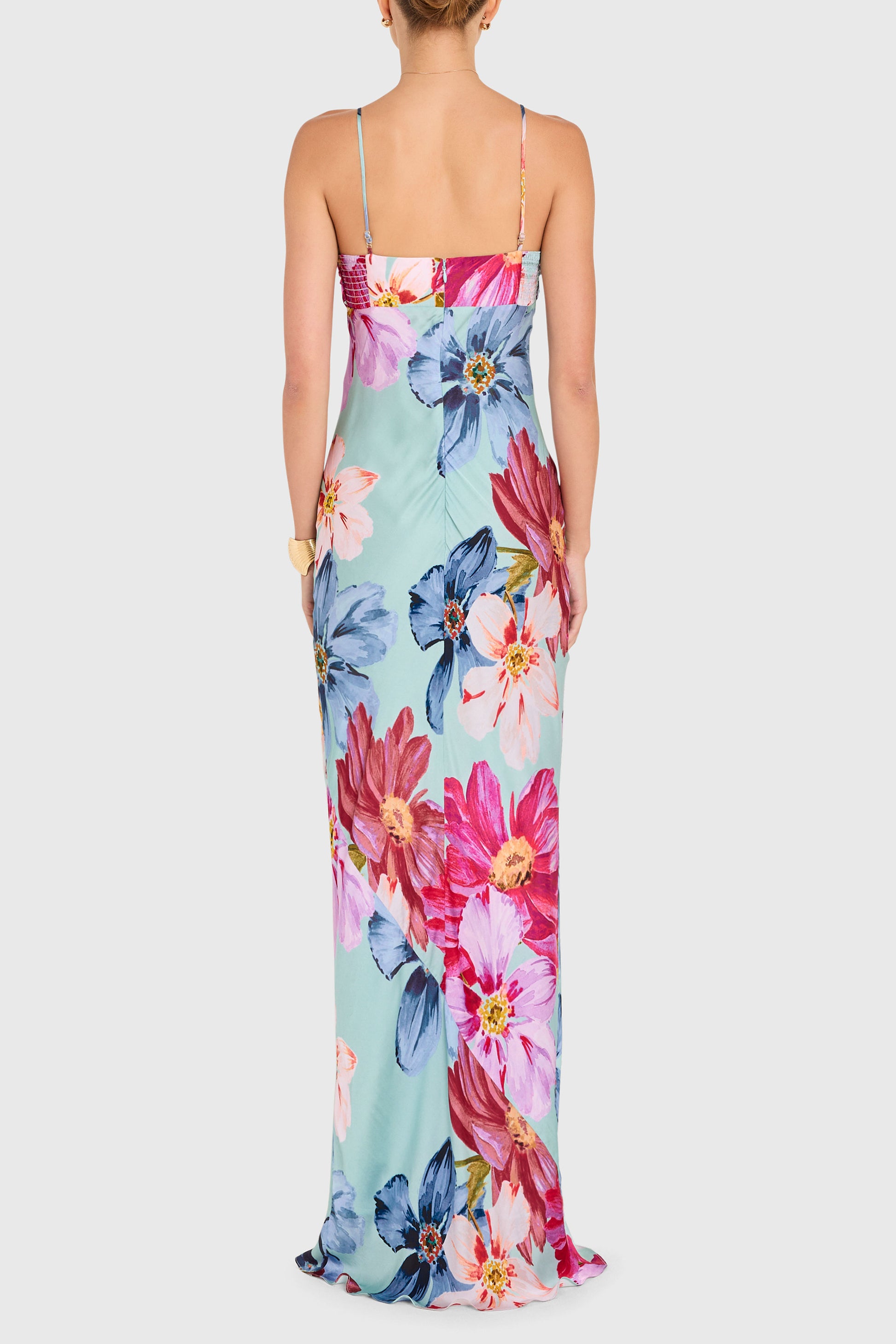 A woman is seen from the back wearing the Amanda Uprichard Ira Maxi Dress—a long, light blue dress with spaghetti straps and large pink, blue, and red floral prints, her hair up as she stands against a plain background.