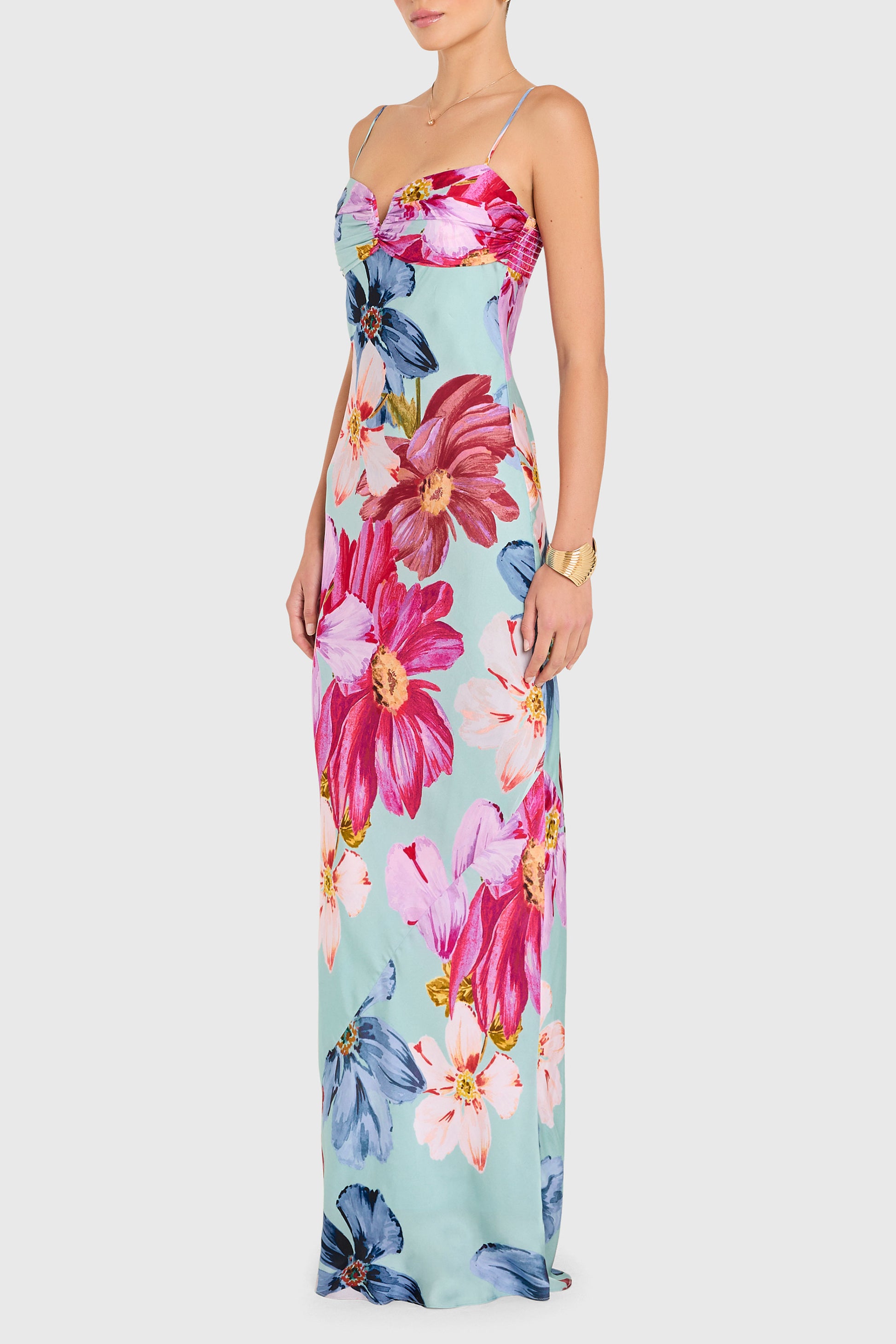 A woman wears the Amanda Uprichard Ira Maxi Dress, a sleeveless piece with spaghetti straps, sweetheart neckline, and bold pink, red, and blue floral print on light blue. She completes the look with a gold bracelet.