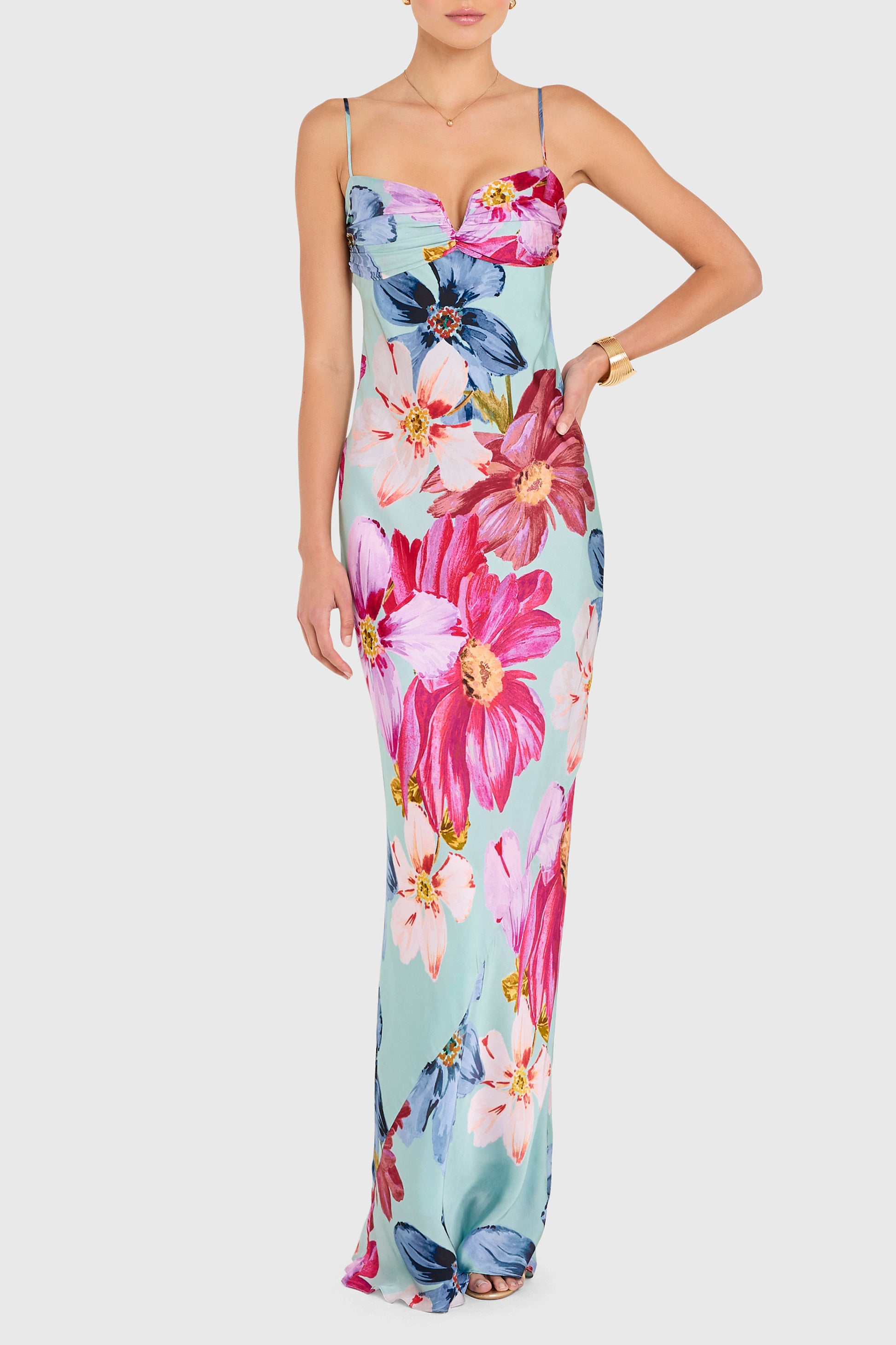 A woman wears the Ira Maxi Dress by Amanda Uprichard— a long, light blue gown featuring spaghetti straps and a bold floral print in pink, red, blue, and white. She poses elegantly with one arm bent and the other at her side.