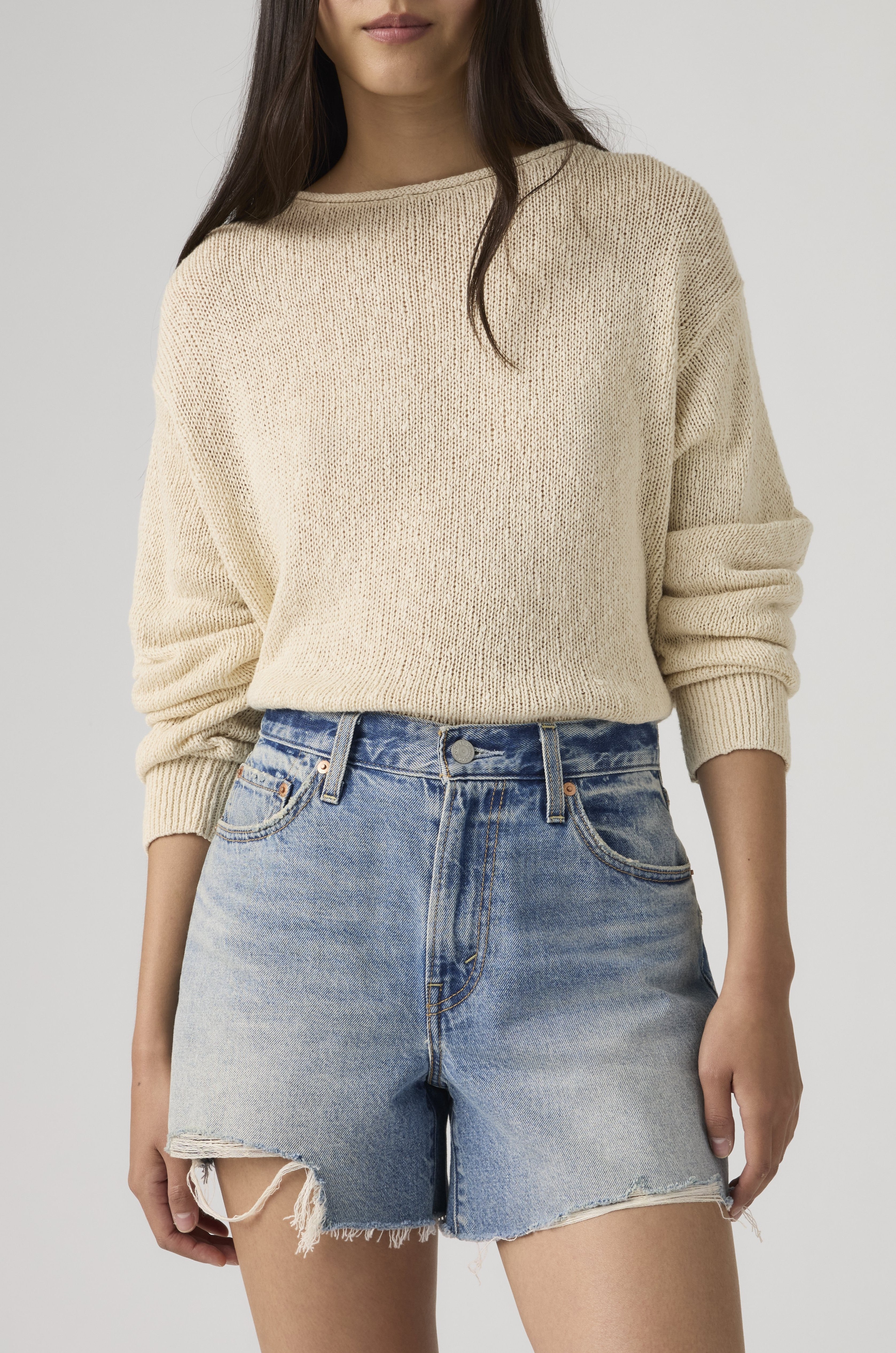 Someone wears a light beige 100% cotton knit sweater with sleeves pushed up and Levi’s Ribcage Short Zip Blow My Mind in light-wash denim with frayed hems, standing against a plain background.