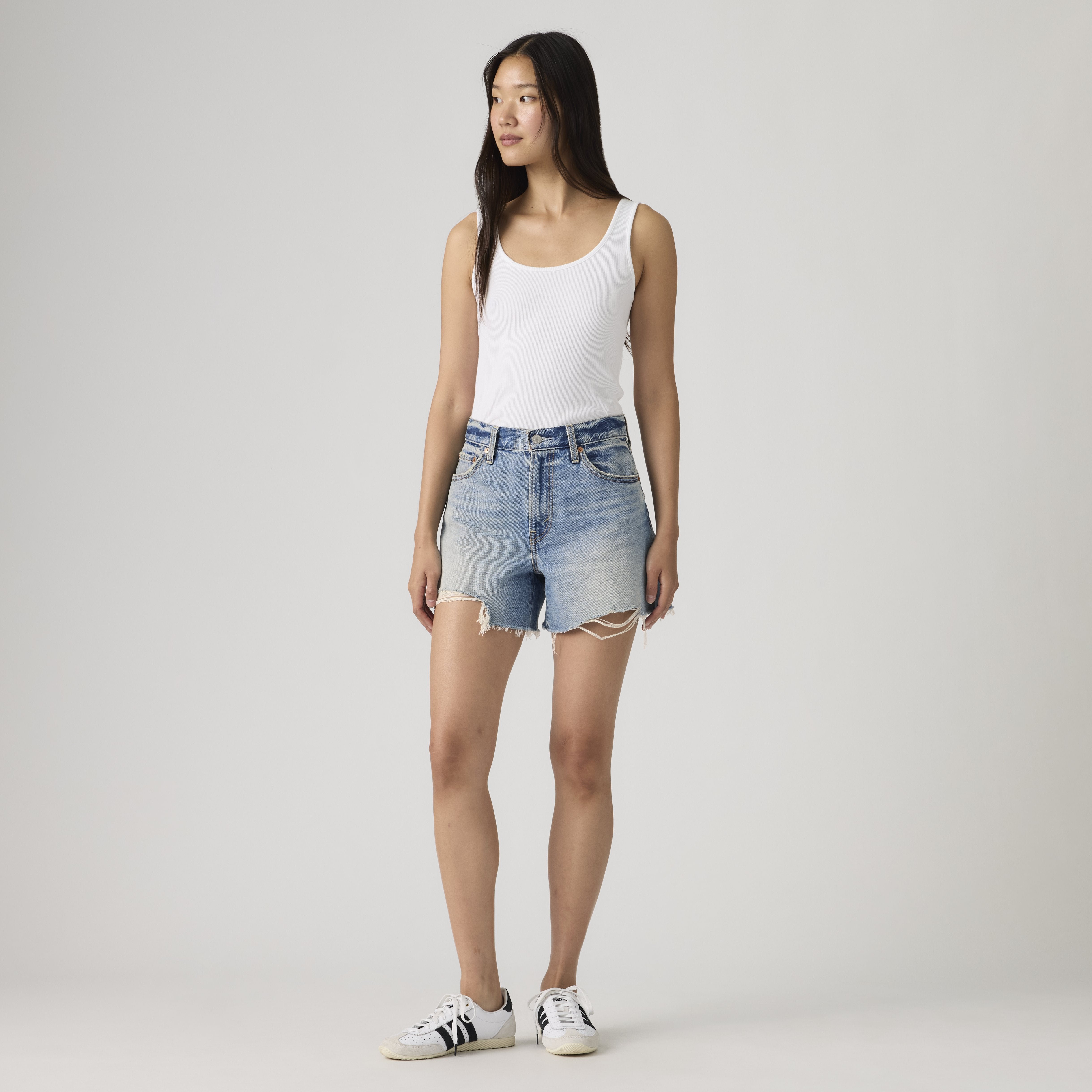 A woman stands against a plain background in a white tank top, Levi’s Ribcage Short Zip Blow My Mind shorts with frayed hems, and white sneakers with black accents, looking slightly to the side.