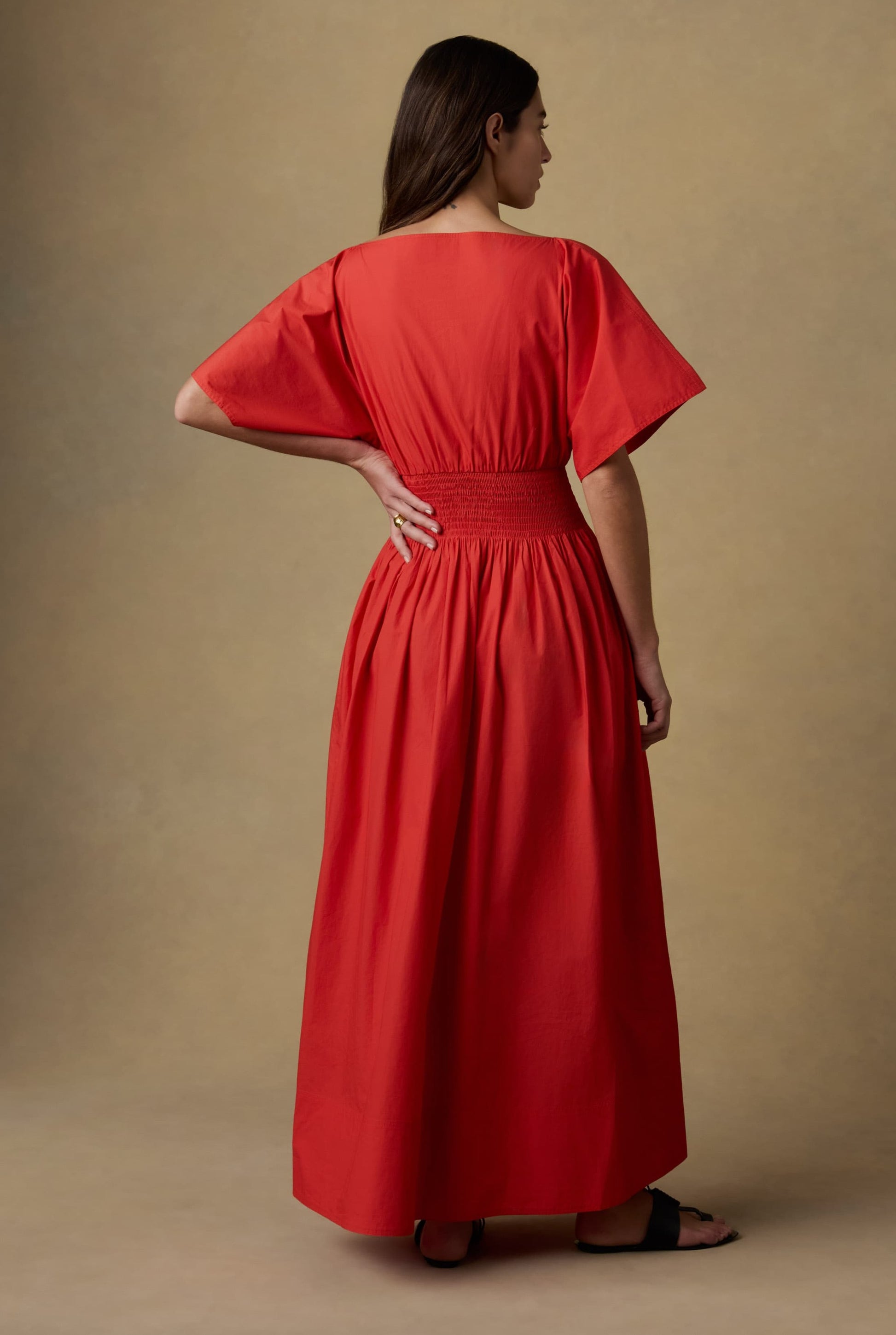 A woman with long brown hair stands with one hand on her hip, facing away, wearing the Faherty Coastal Poplin Elia Smocked Dress in bright red against a neutral background.