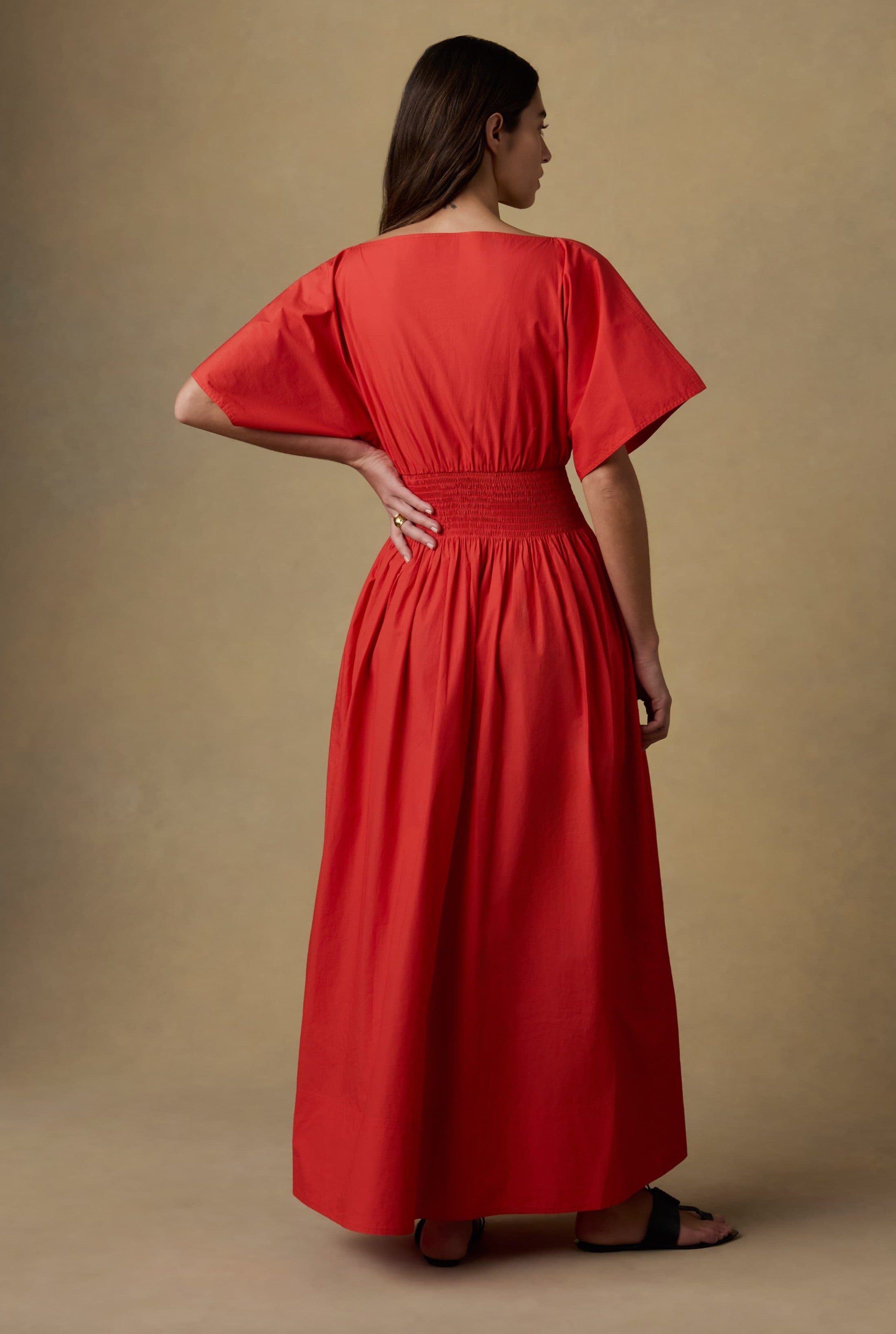 A woman with long brown hair stands with one hand on her hip, facing away, wearing the Faherty Coastal Poplin Elia Smocked Dress in bright red against a neutral background.