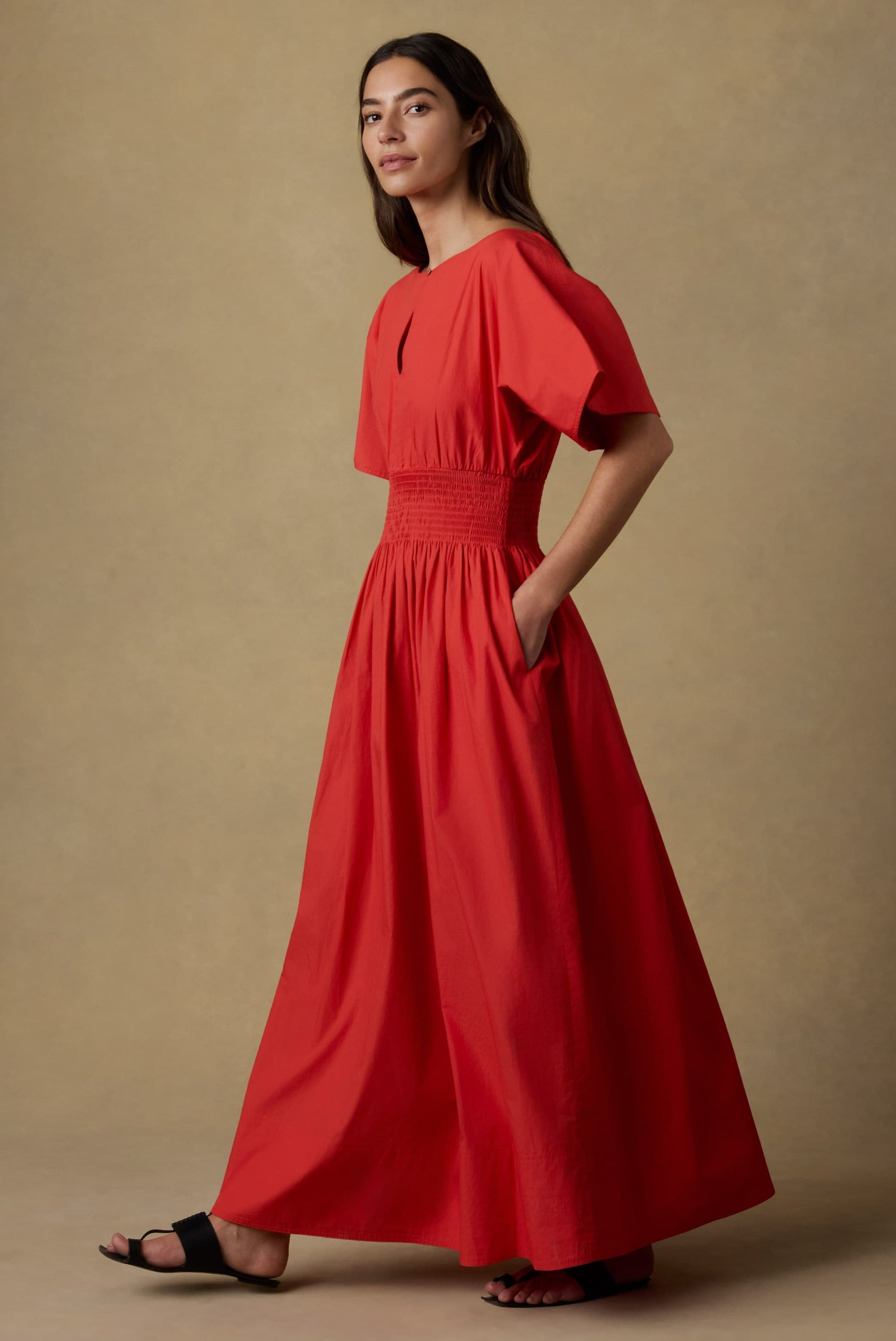 A woman stands sideways in the Faherty Coastal Poplin Elia Smocked Dress, featuring an A-line silhouette, short sleeves, smocked waist, and side pockets. She has long dark hair and wears black sandals against a neutral backdrop.
