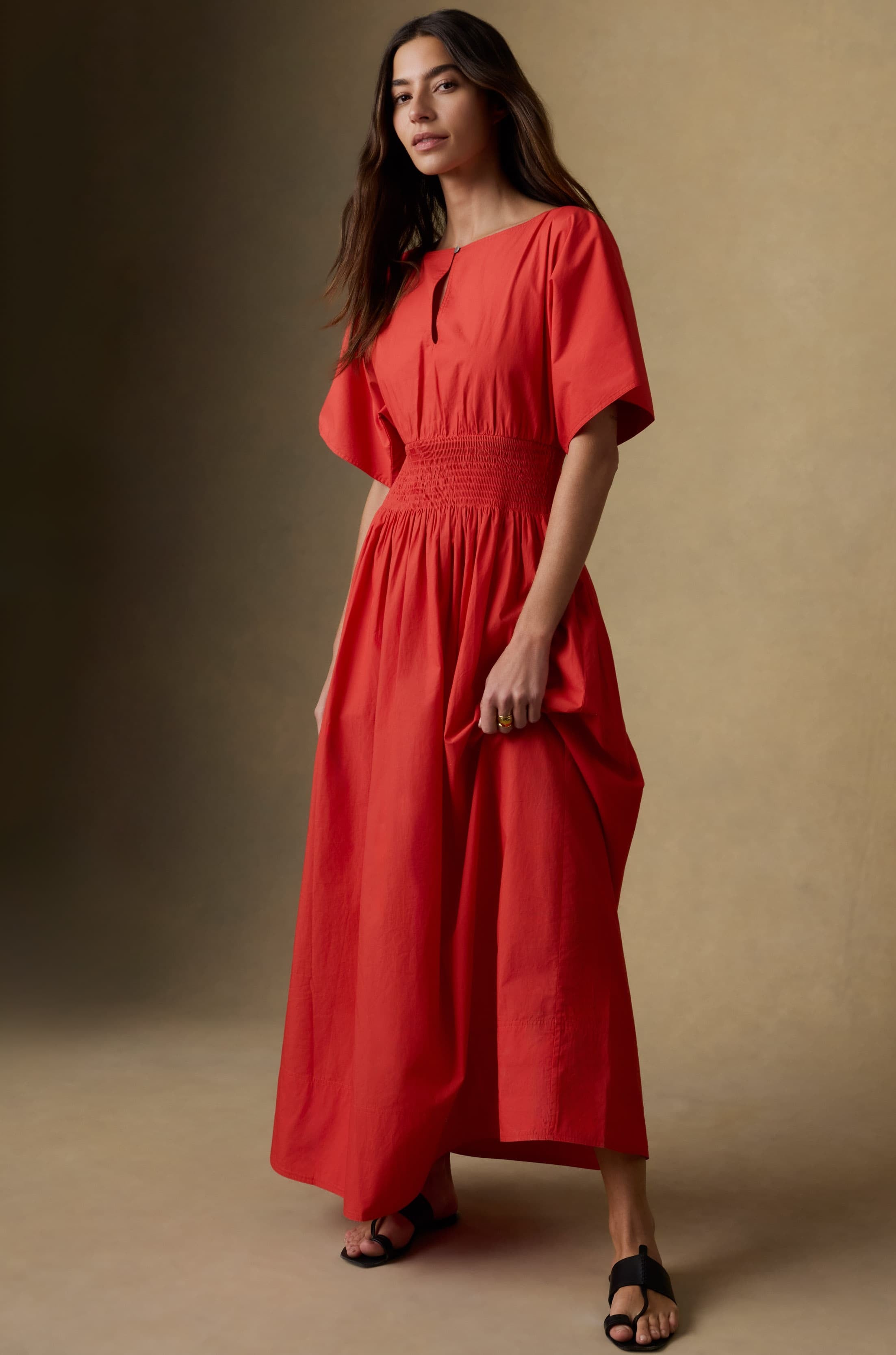 A woman with long, wavy hair stands against a neutral background wearing the Faherty Coastal Poplin Elia Smocked Dress—an A-line, bright red cotton maxi dress with short sleeves, a cinched waist, and black sandals.